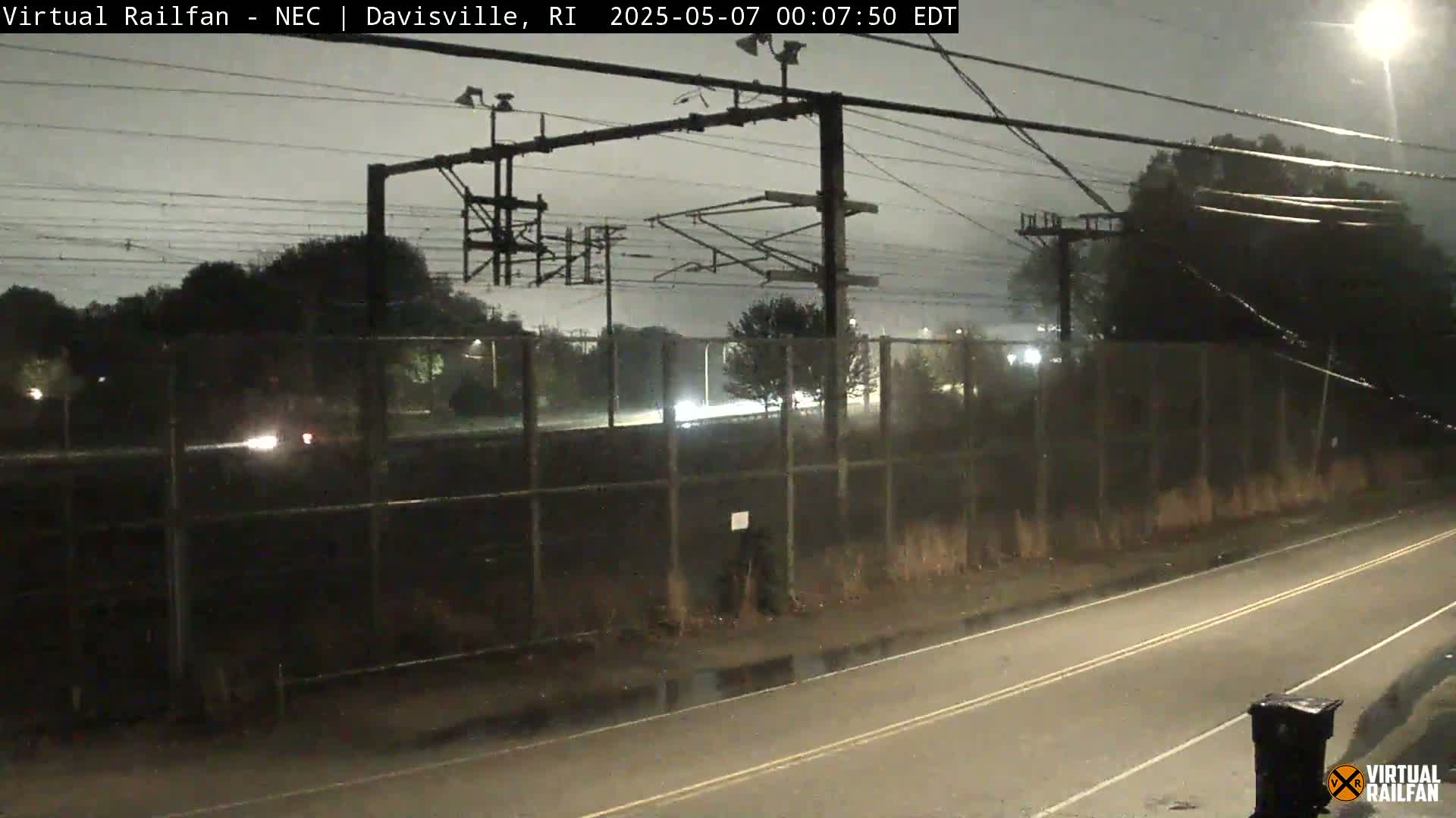 A nighttime view of a road next to a chain-link fence separating it from a railway under a dark, overcast sky.