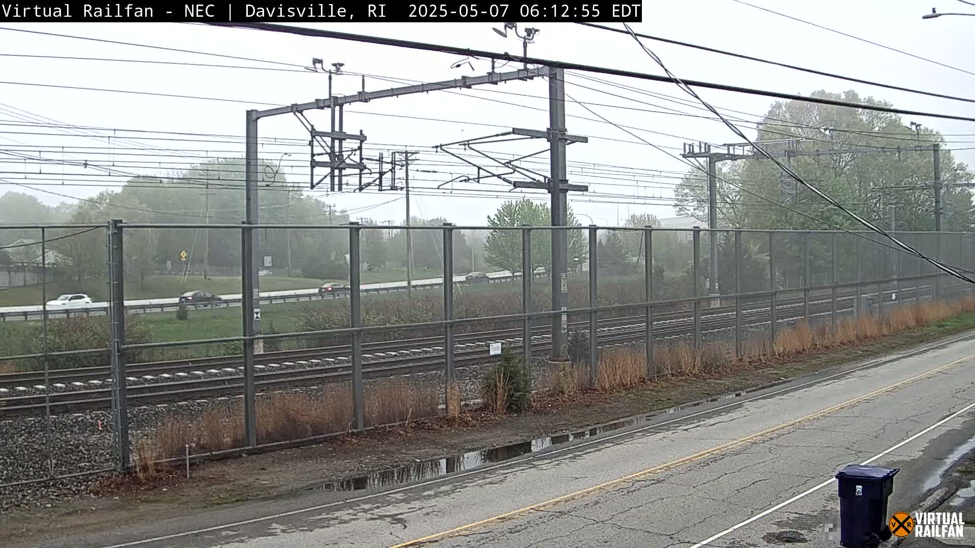 A rain-misted view of railroad tracks behind a chain-link fence, alongside a road with puddles, and some cars driving on a road beyond the fence.