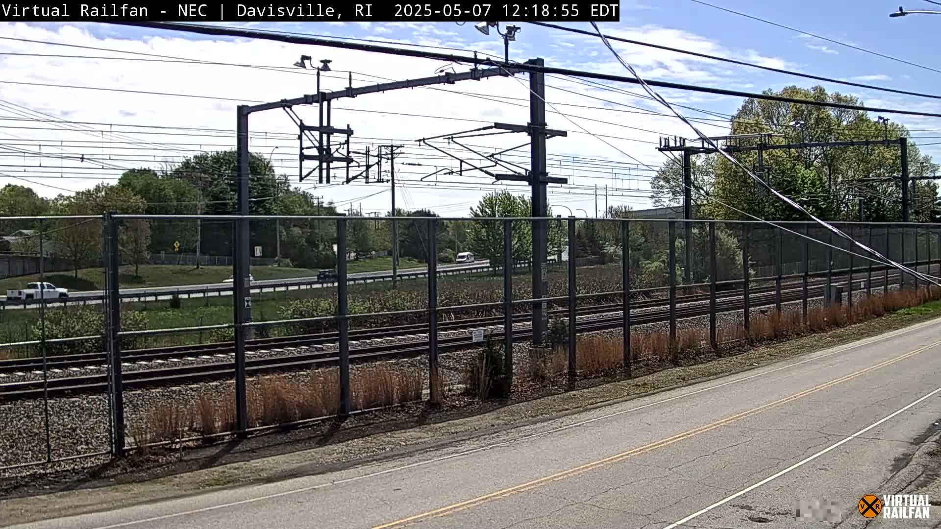 A sunny day shows a view of train tracks alongside a road, separated by a chain link fence, with trees and cars visible in the background.