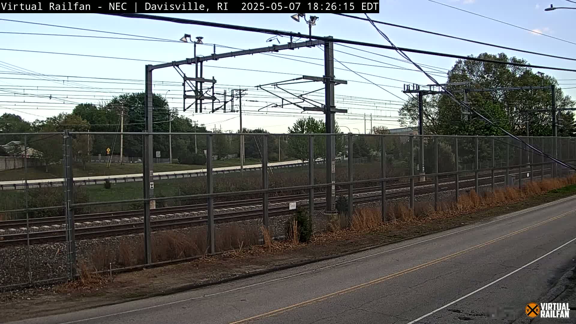 A view of railroad tracks behind a chain-link fence beside a road on a partly cloudy day.