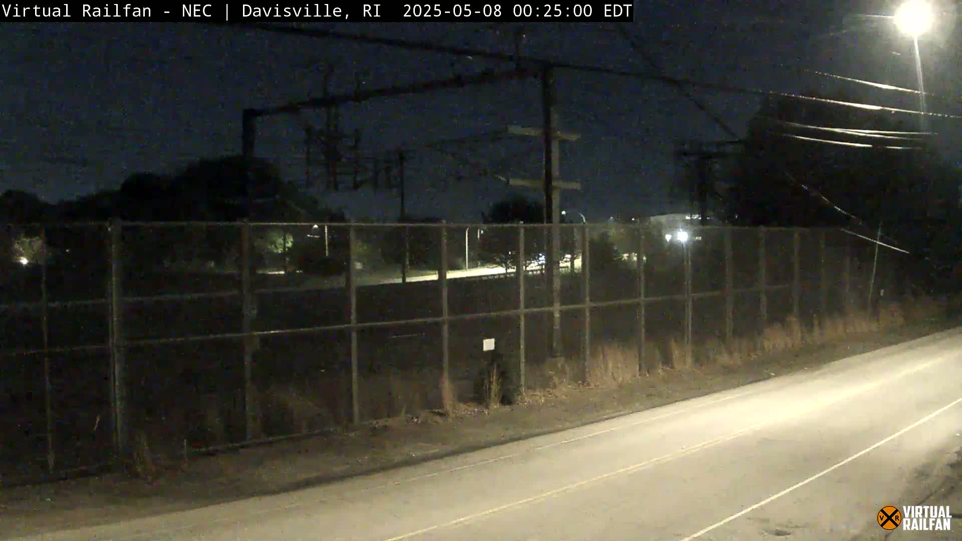 A nighttime view of a road next to a chain-link fence, beyond which are dimly lit trees and buildings under a clear, dark sky.