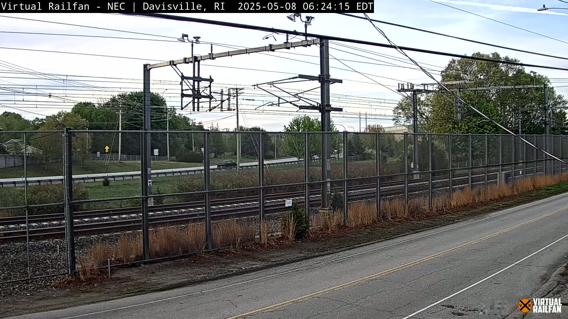 A view of railroad tracks bordered by a chain-link fence, with a road running alongside, under a partly cloudy sky.