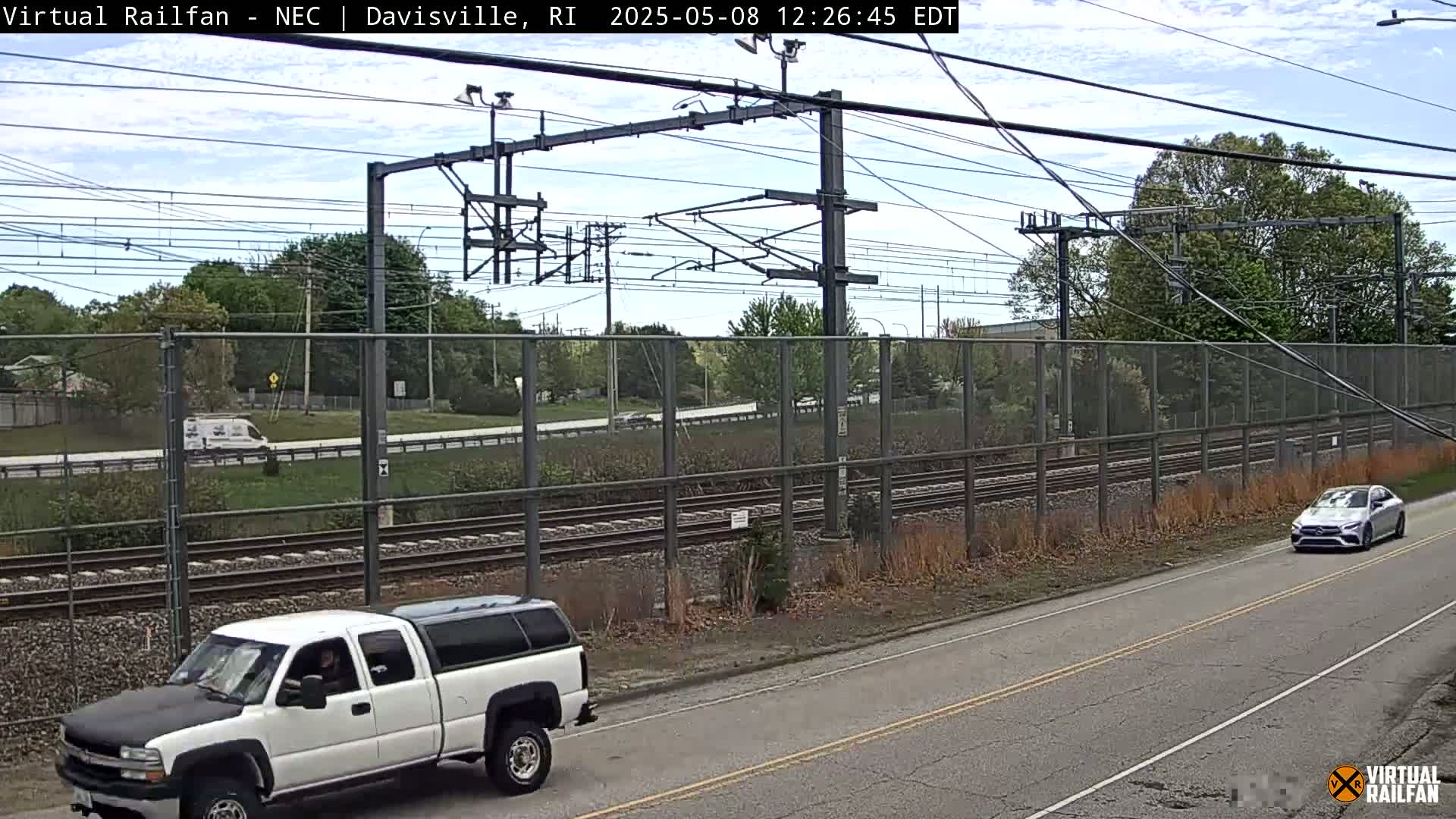 A partly cloudy day shows a white pickup truck and a silver car parked on a road next to railroad tracks and a chain-link fence.