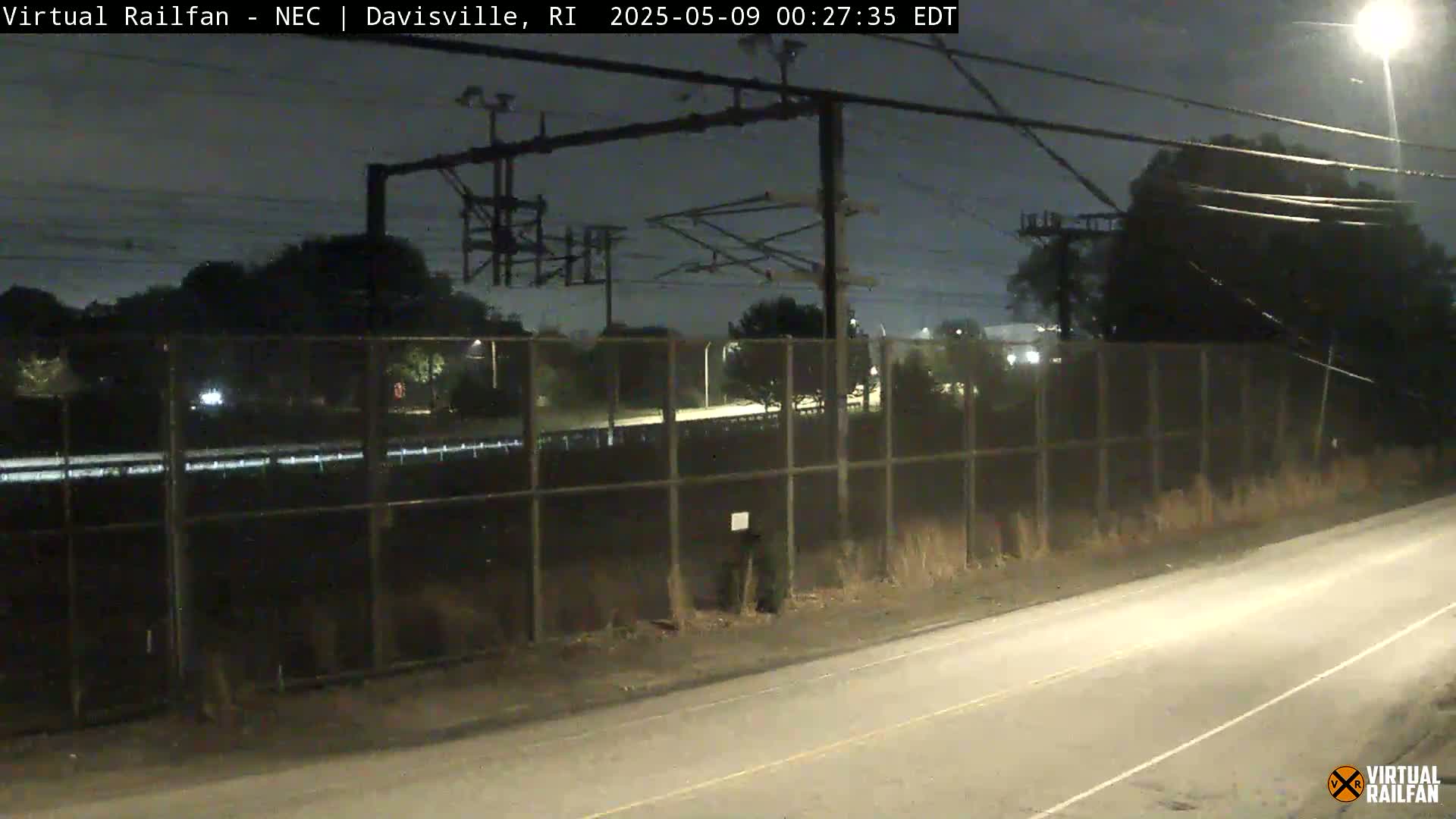 A nighttime view of a road beside a chain-link fence, beyond which are trees and overhead power lines.