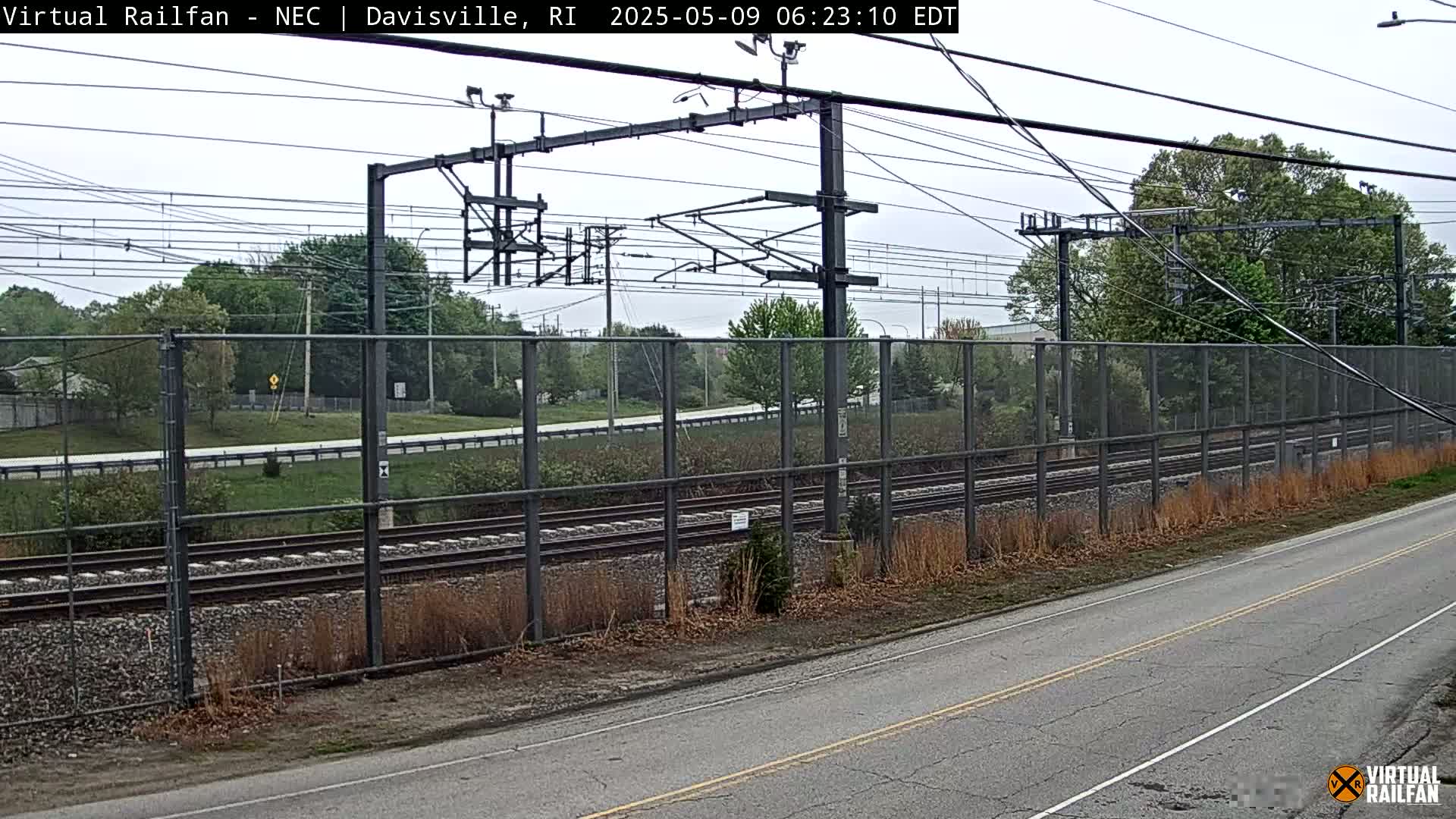 A view of train tracks behind a chain-link fence, alongside a road, on an overcast day.