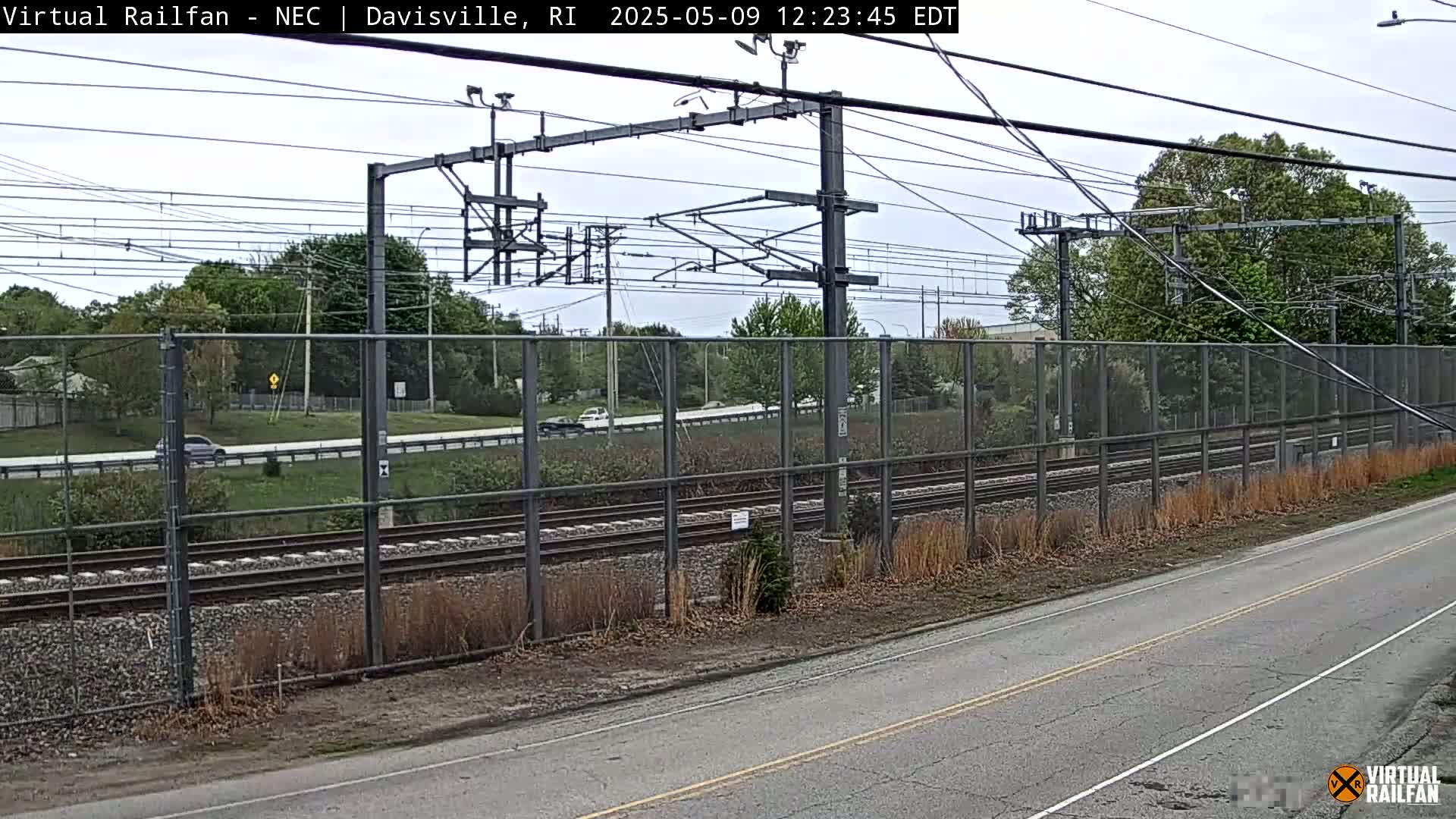 A view of a roadway next to a chain-link fence separating it from railroad tracks under an overcast sky.