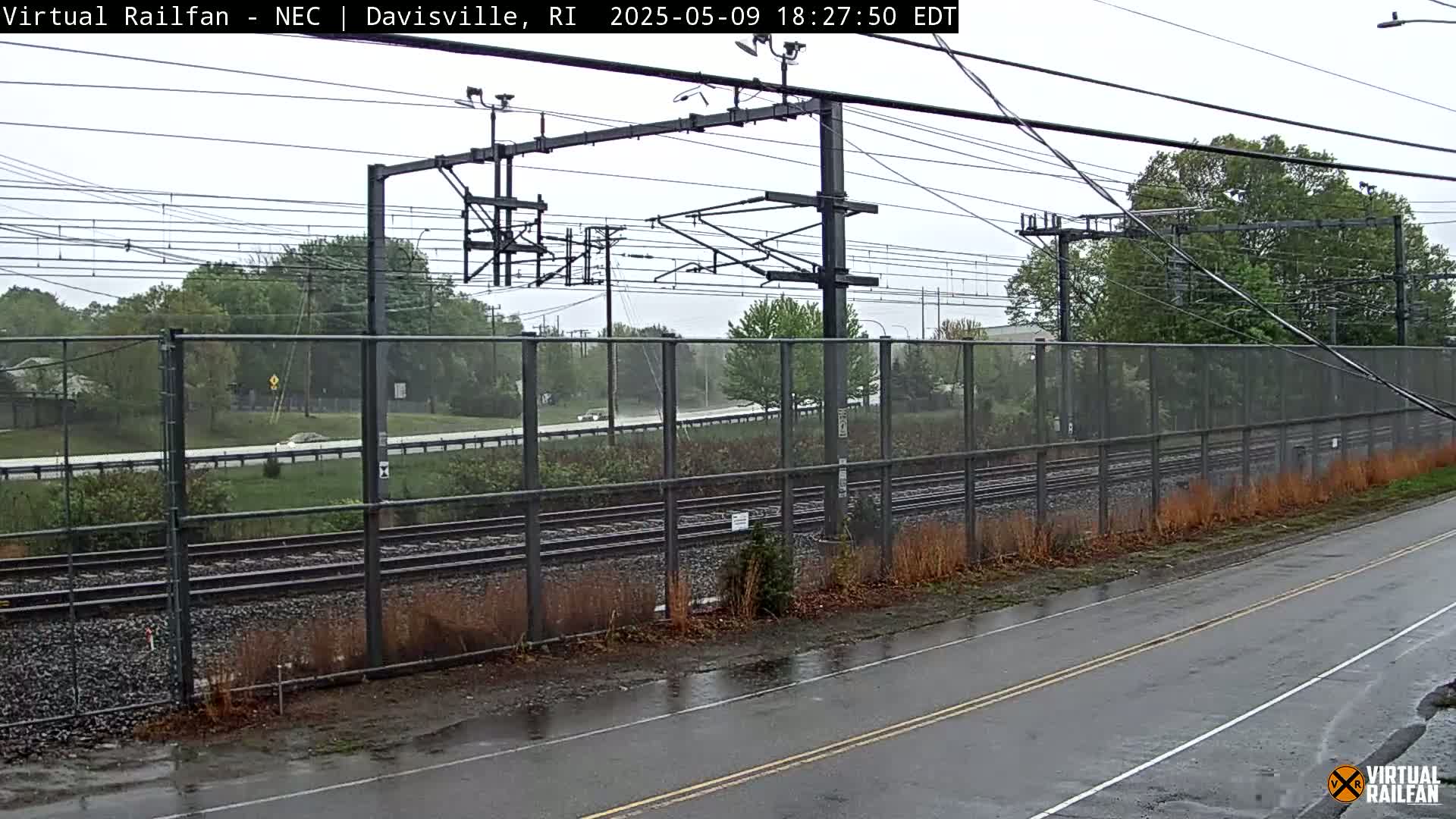 A wet road next to a chain-link fence runs alongside railroad tracks under an overcast sky.