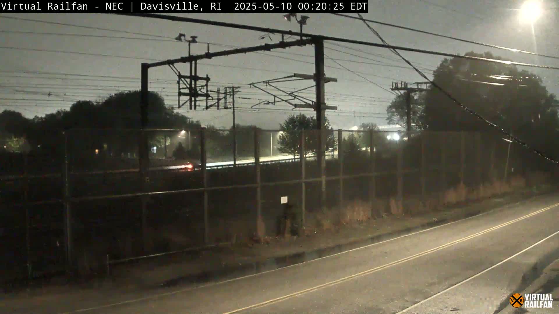 A nighttime view of a rain-slicked road beside a chain-link fence, beyond which are railway lines and trees under a dark, overcast sky.