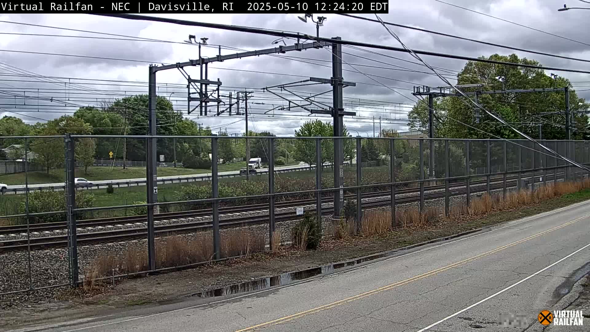 A partly cloudy day shows a view of train tracks beside a road, with cars and a truck visible beyond a chain-link fence.