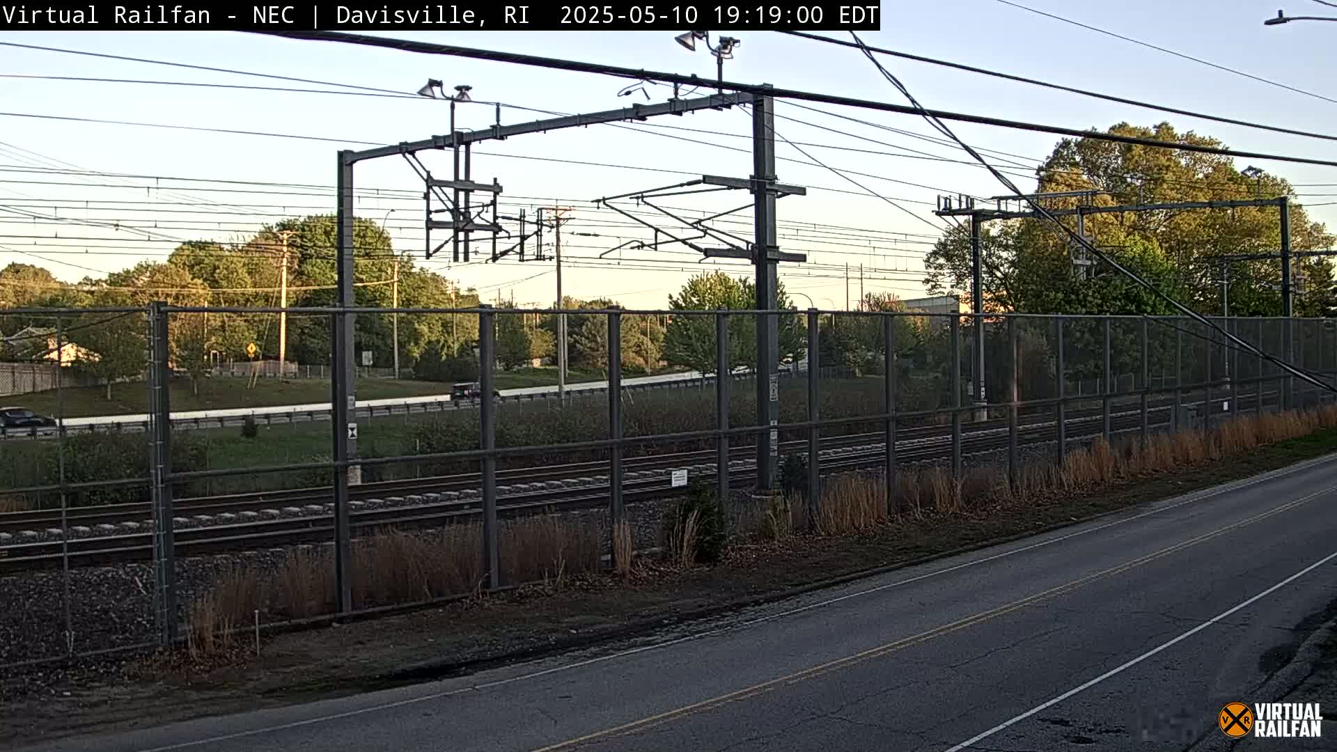 A view of railroad tracks beside a road, separated by a chain-link fence, under a clear evening sky.