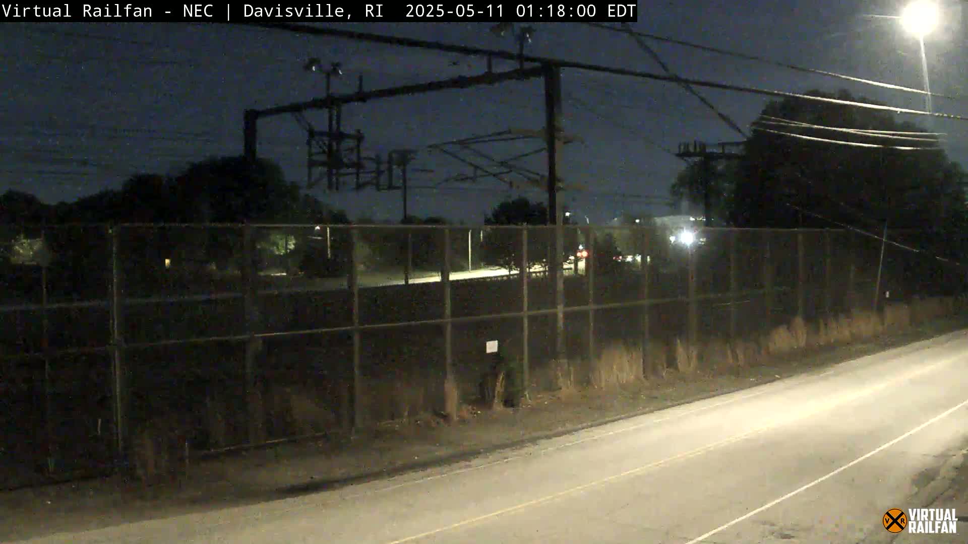A nighttime view of a road beside a chain-link fence, beyond which are train tracks and overhead electrical lines under a dark, clear sky.