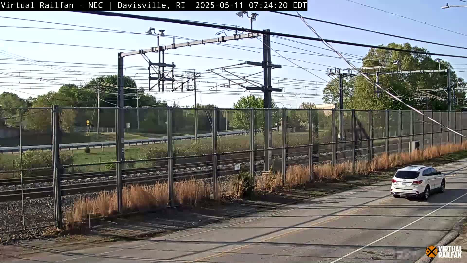 A white SUV drives on a road next to a chain-link fence separating it from railroad tracks and a grassy area under a clear sky.