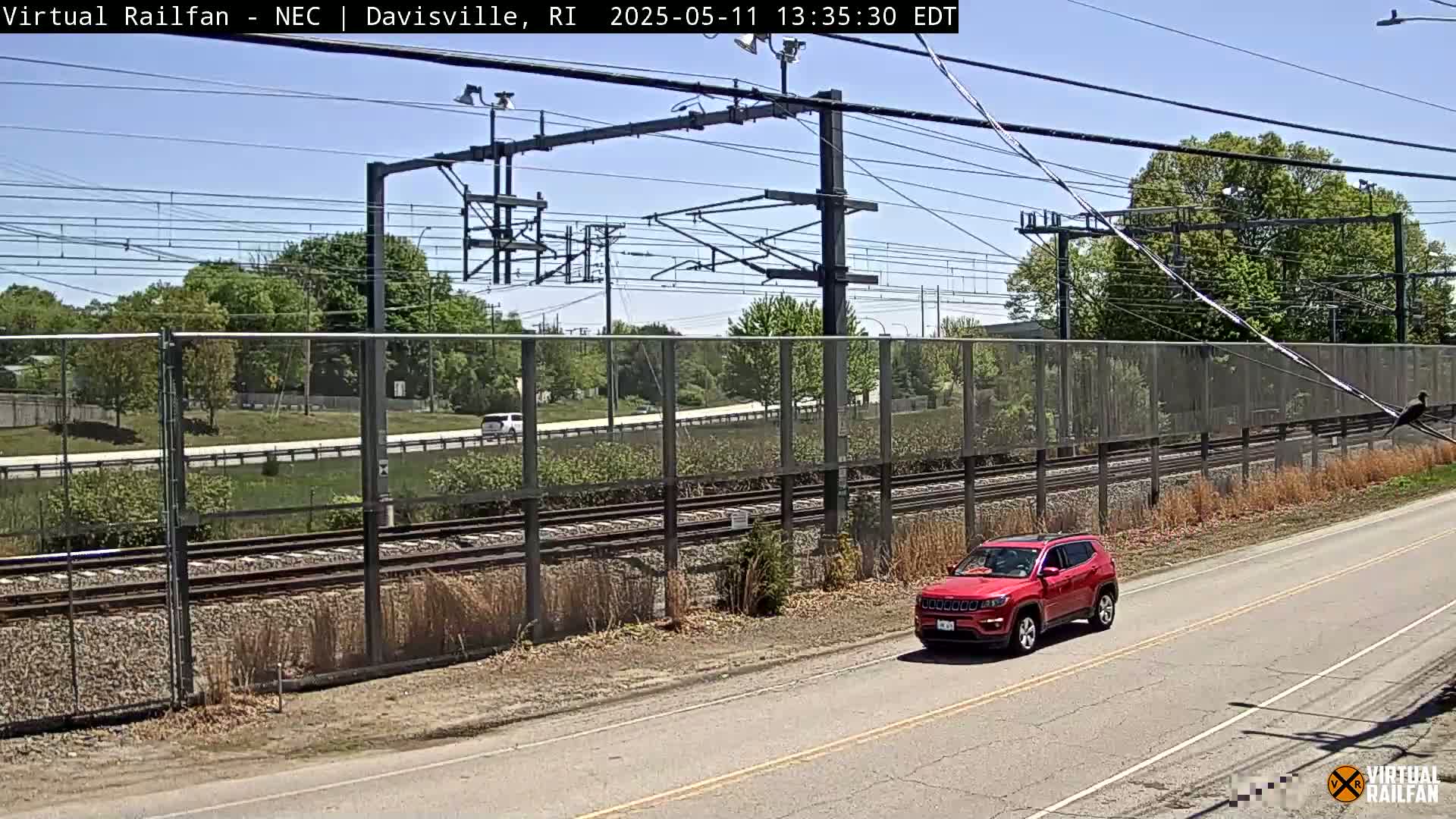 A red SUV drives on a road next to a railway track bordered by a fence on a sunny day.