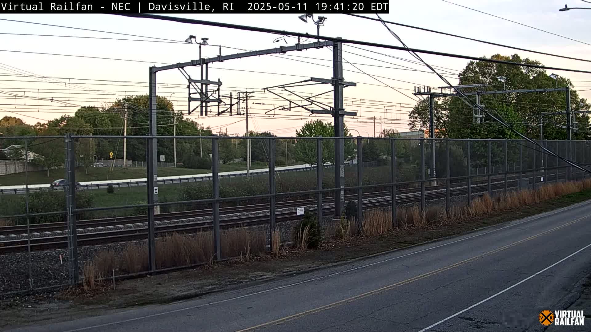 A view of railroad tracks beside a road, fenced in by a chain link fence, under a partly cloudy, twilight sky.
