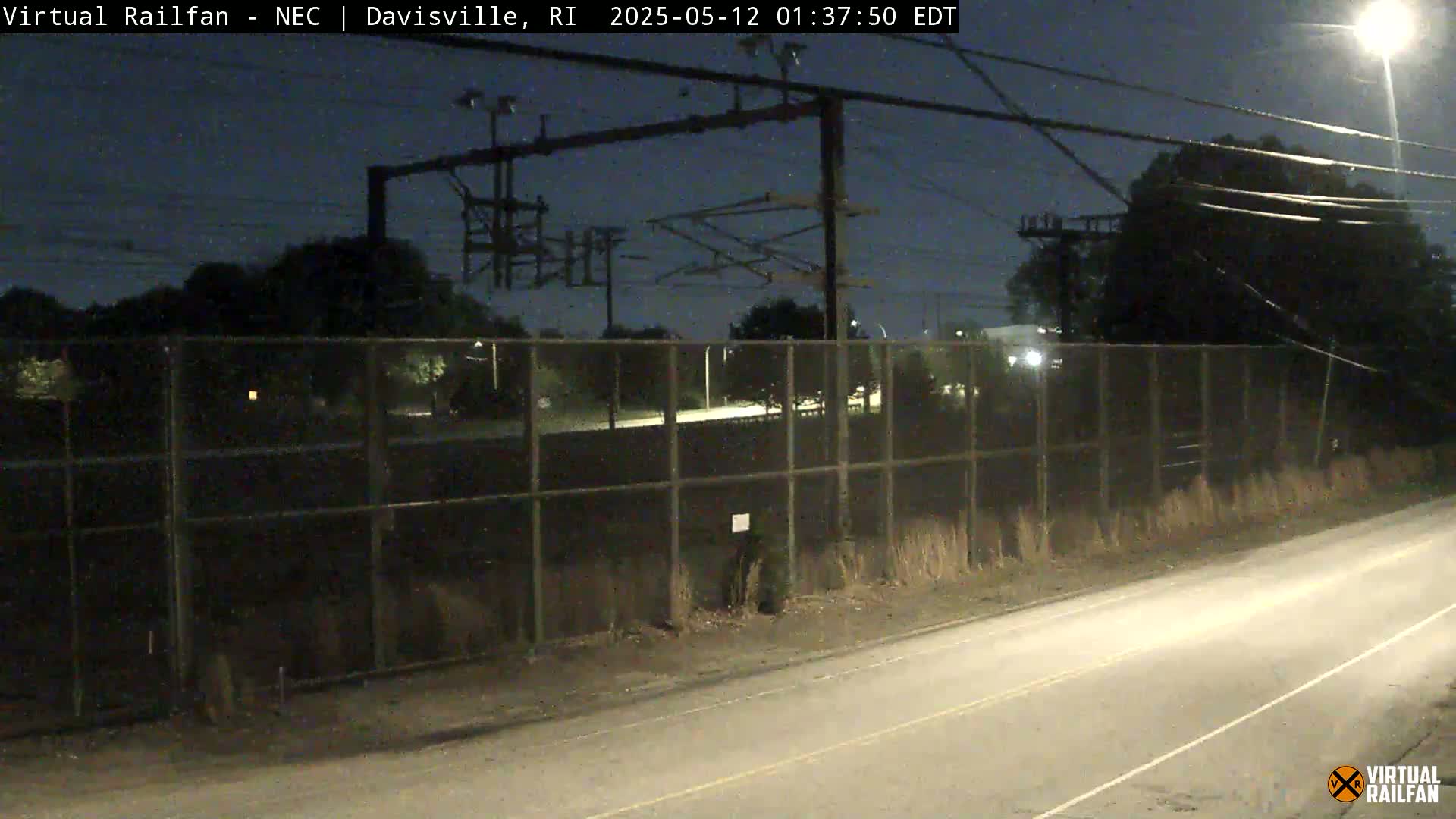 A nighttime view of a chain-link fence bordering a road, with overhead power lines and trees beyond the fence under a dark sky.