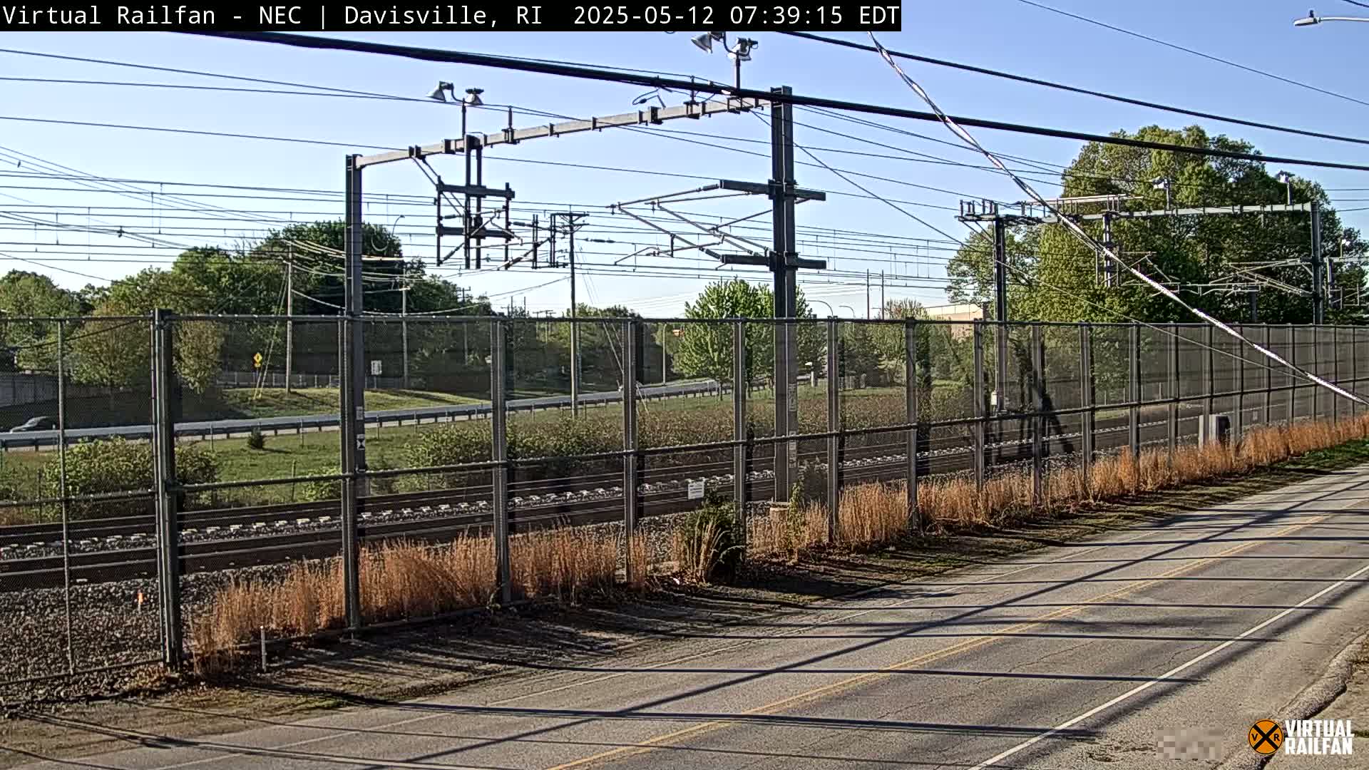 A sunny view of a chain-link fence separating a paved road from railroad tracks and a grassy area beyond.