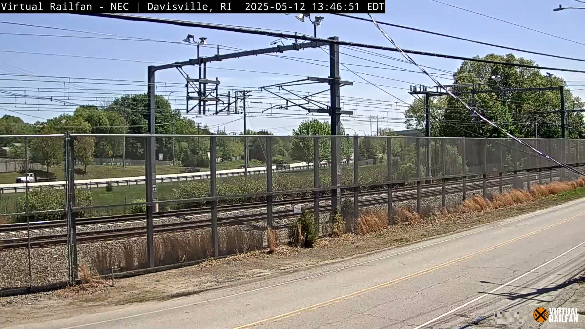 A sunny day view of train tracks behind a chain link fence, beside a road with green trees and grass in the background.