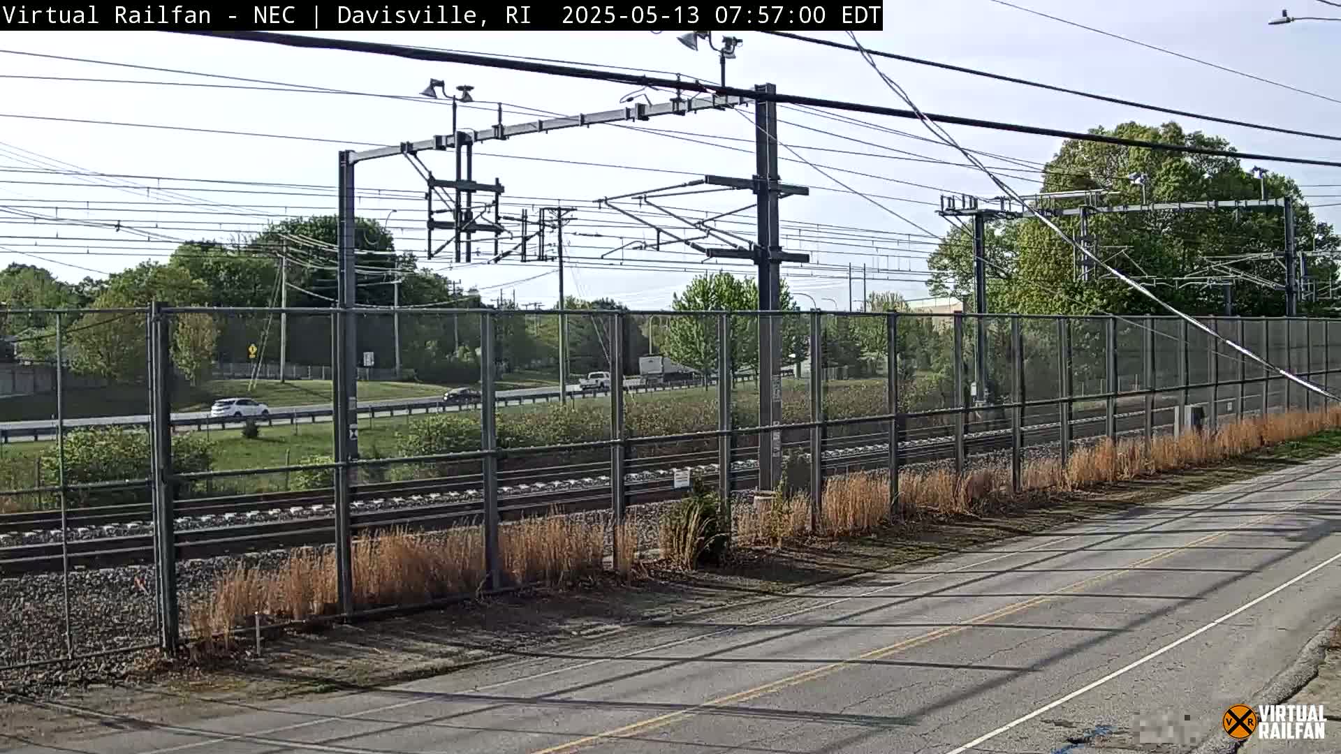 A view of a roadway and railroad tracks beside a chain-link fence, with cars on the road and trees and shrubs in the background on a partly sunny day.