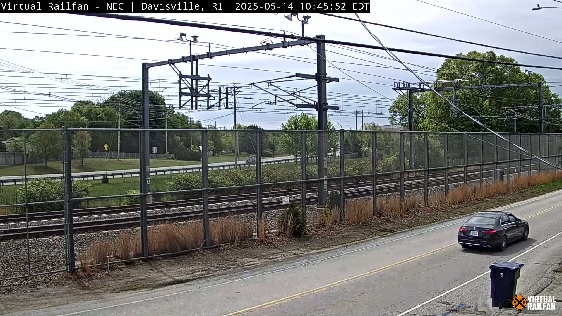 A dark-colored sedan drives on a road next to a chain-link fence bordering a railway under a cloudy sky.