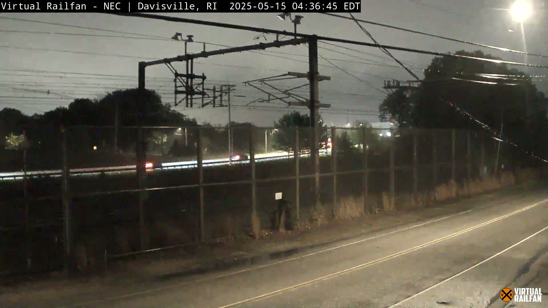 A nighttime view of a highway and train tracks separated by a fence, under a dark, possibly rainy sky.