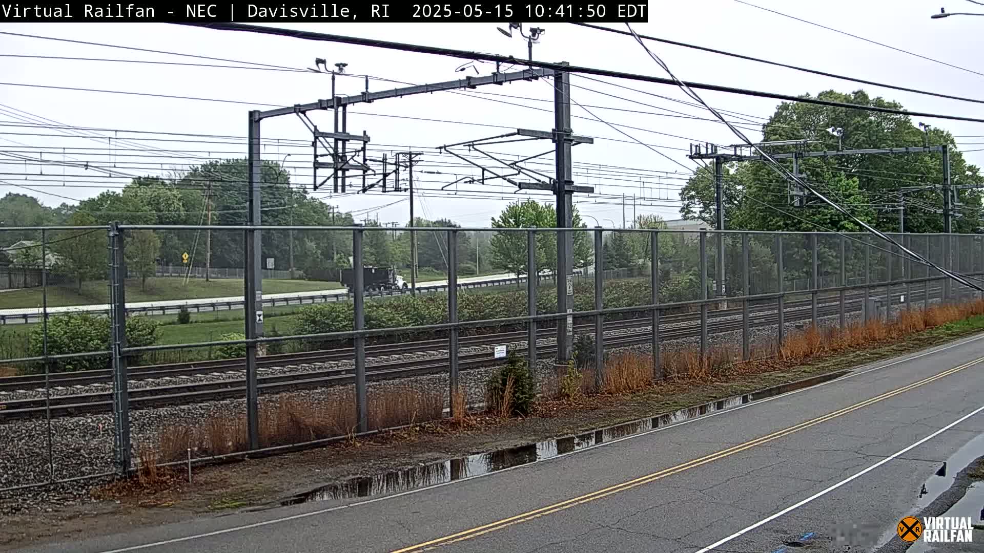 A view of railroad tracks beside a road with a chain-link fence separating them, under an overcast sky with some puddles on the road.