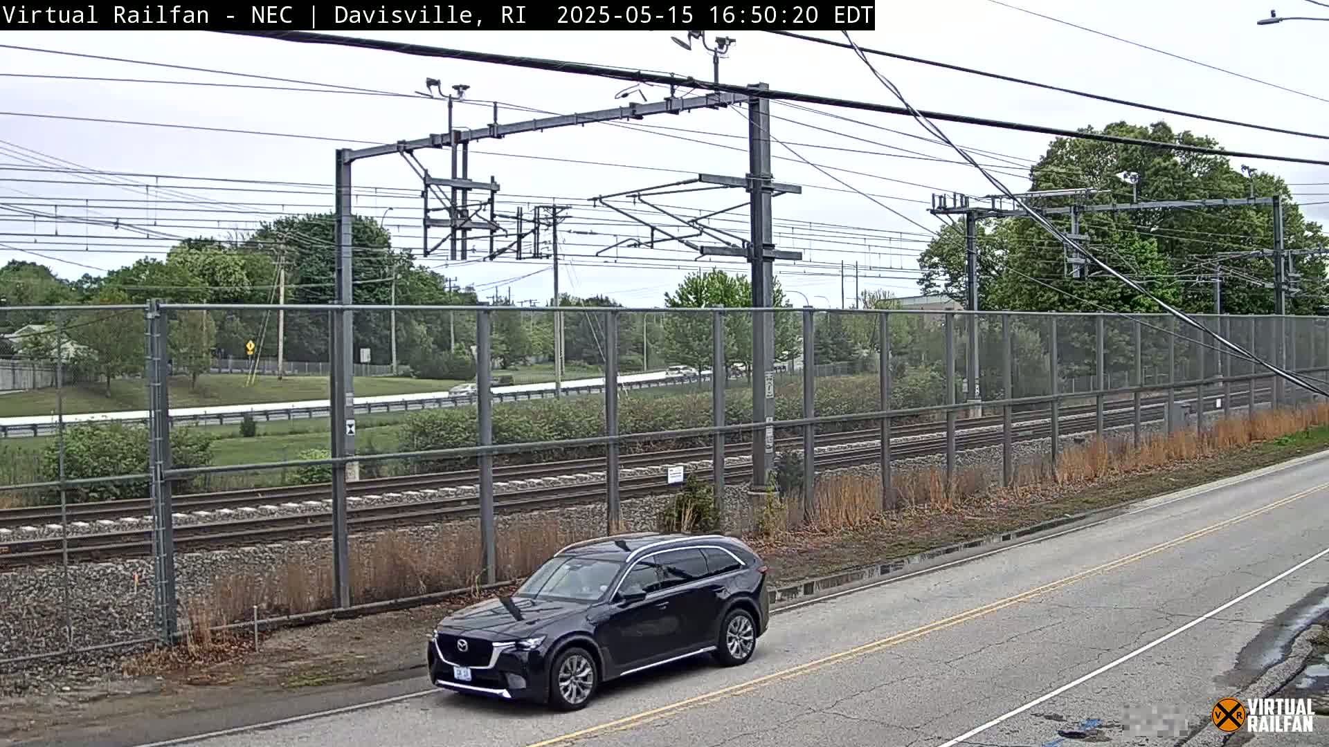 A dark-colored SUV is parked on a road next to a chain-link fence bordering a set of train tracks under an overcast sky.