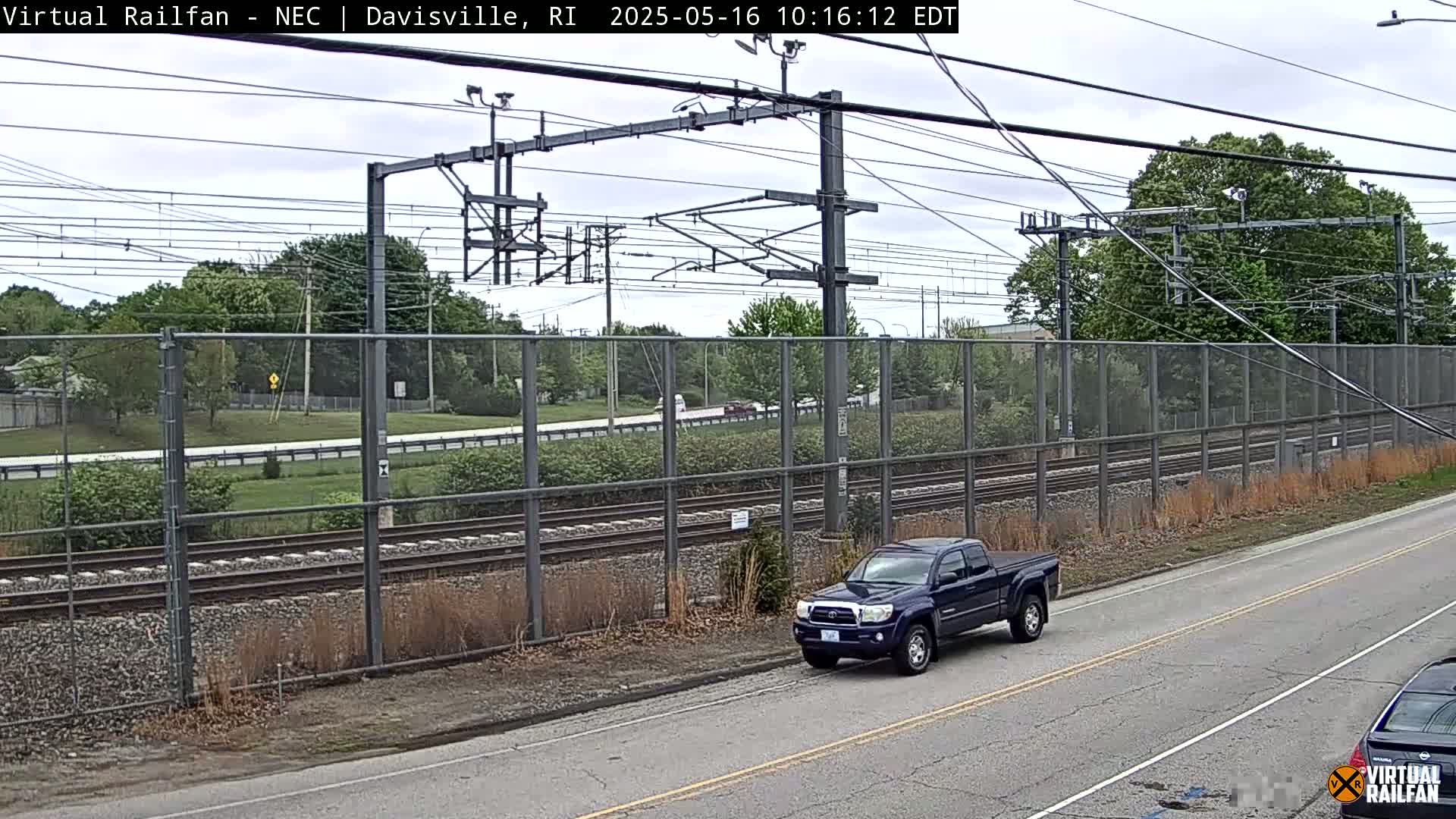 A dark-blue pickup truck is parked on the side of a road next to a chain-link fence separating the road from a railway line under a partly cloudy sky.