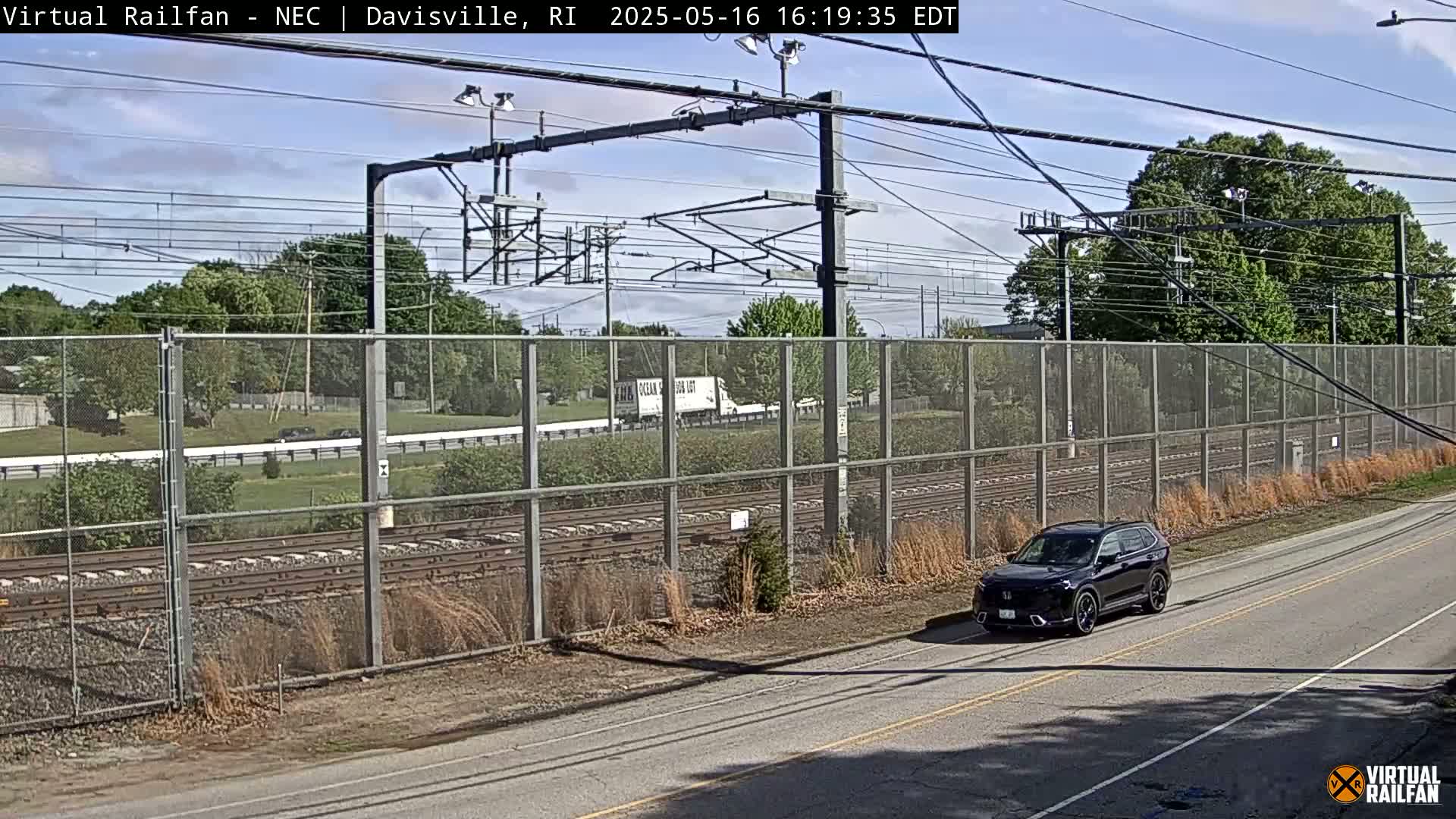 A dark-colored SUV drives on a road next to a chain-link fence bordering railroad tracks under a partly cloudy sky.