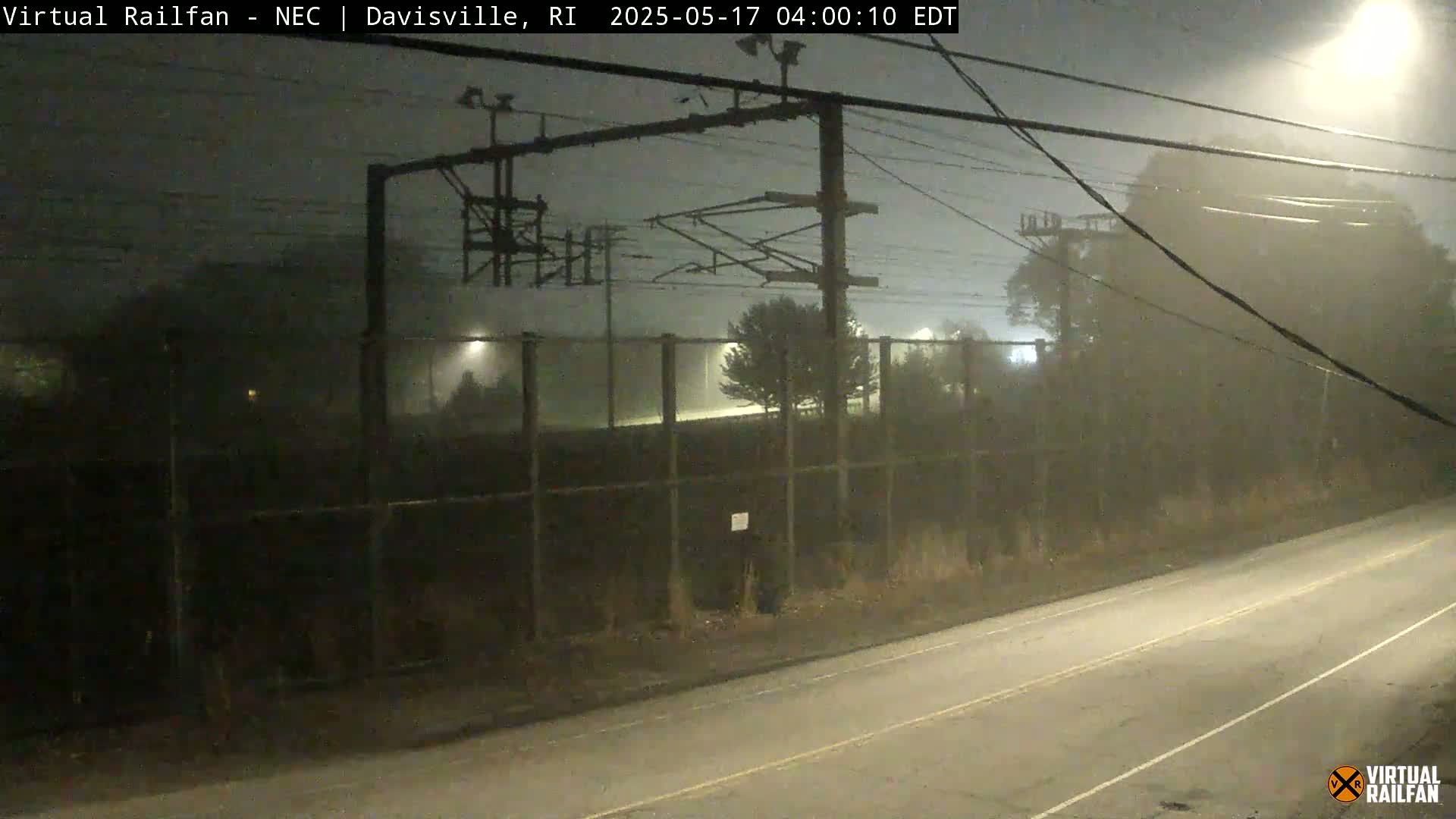 A fog-shrouded nighttime scene shows a chain-link fence alongside a road, with power lines and electrical equipment visible in the background.