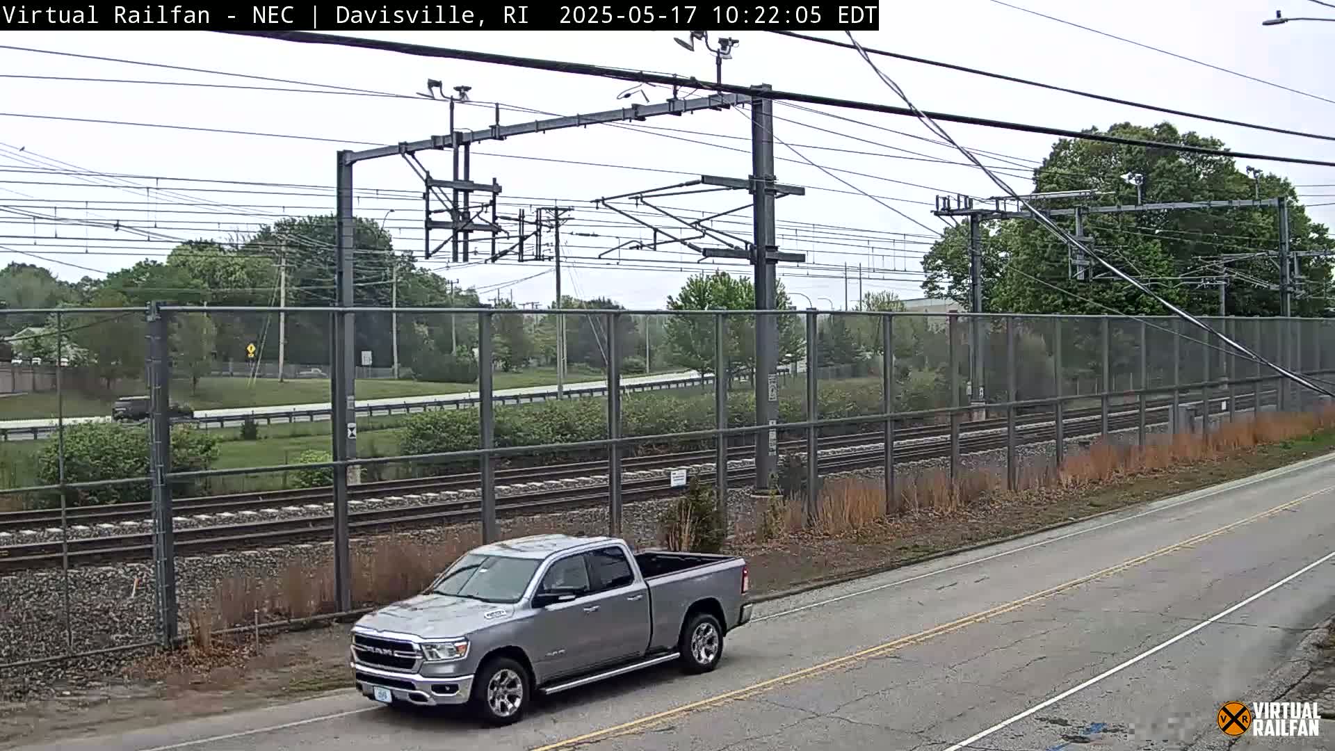 A silver pickup truck is parked on a road next to a chain-link fence separating the road from railroad tracks under an overcast sky.