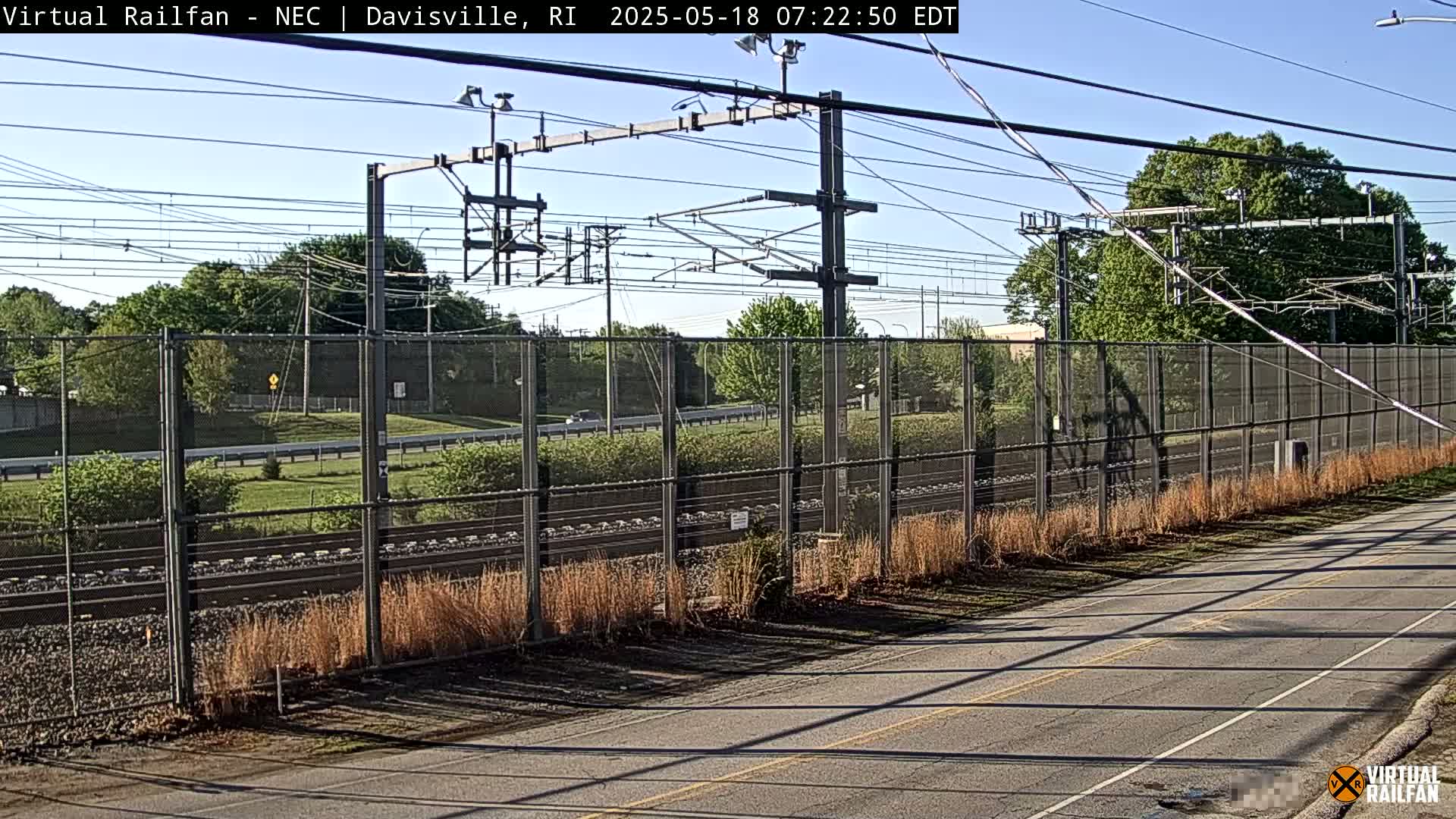 A roadway runs beside a chain-link fence enclosing railroad tracks under a clear, sunny sky.