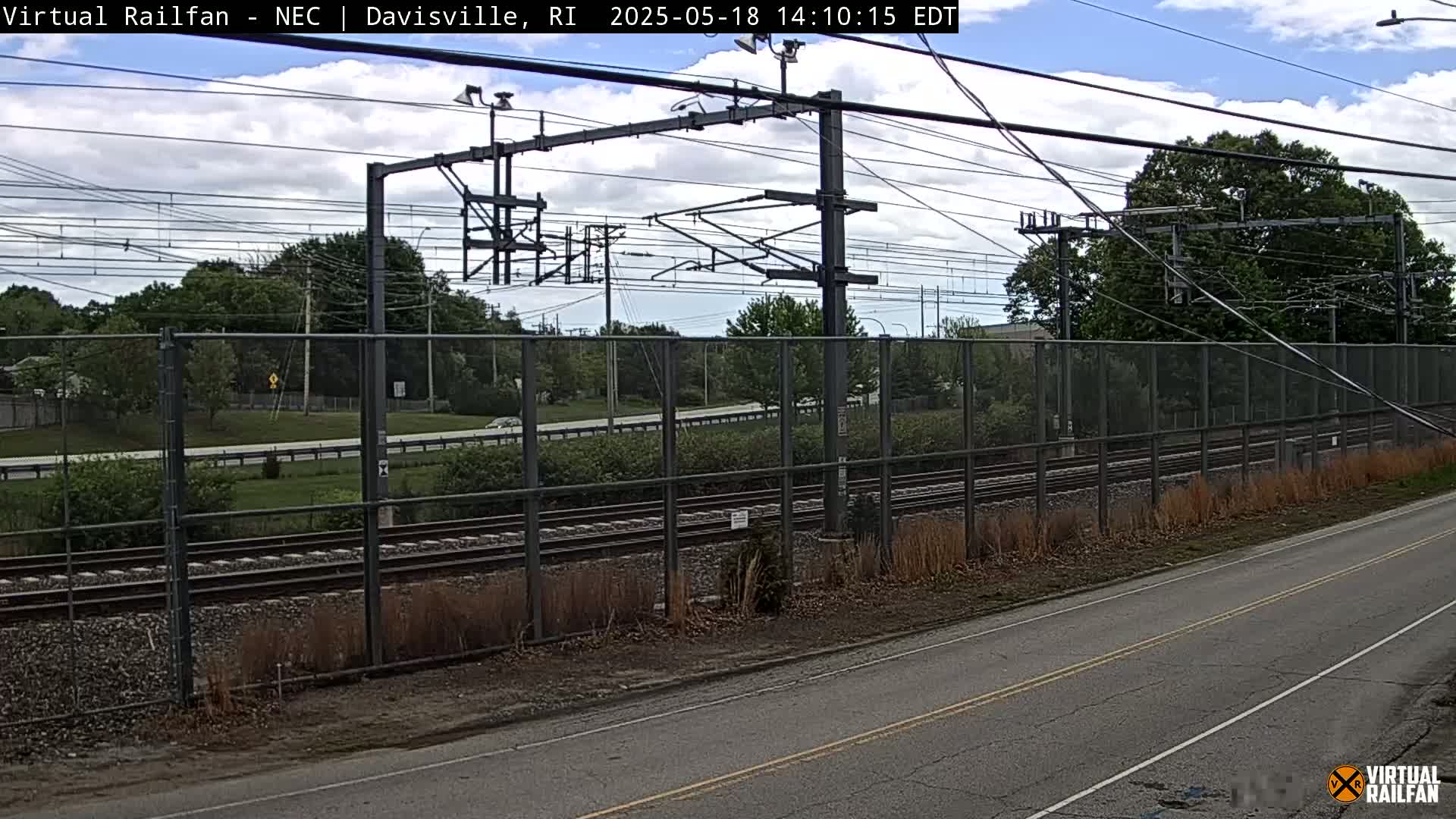 A view of railroad tracks behind a chain-link fence on an overcast day, with a road visible in the foreground.
