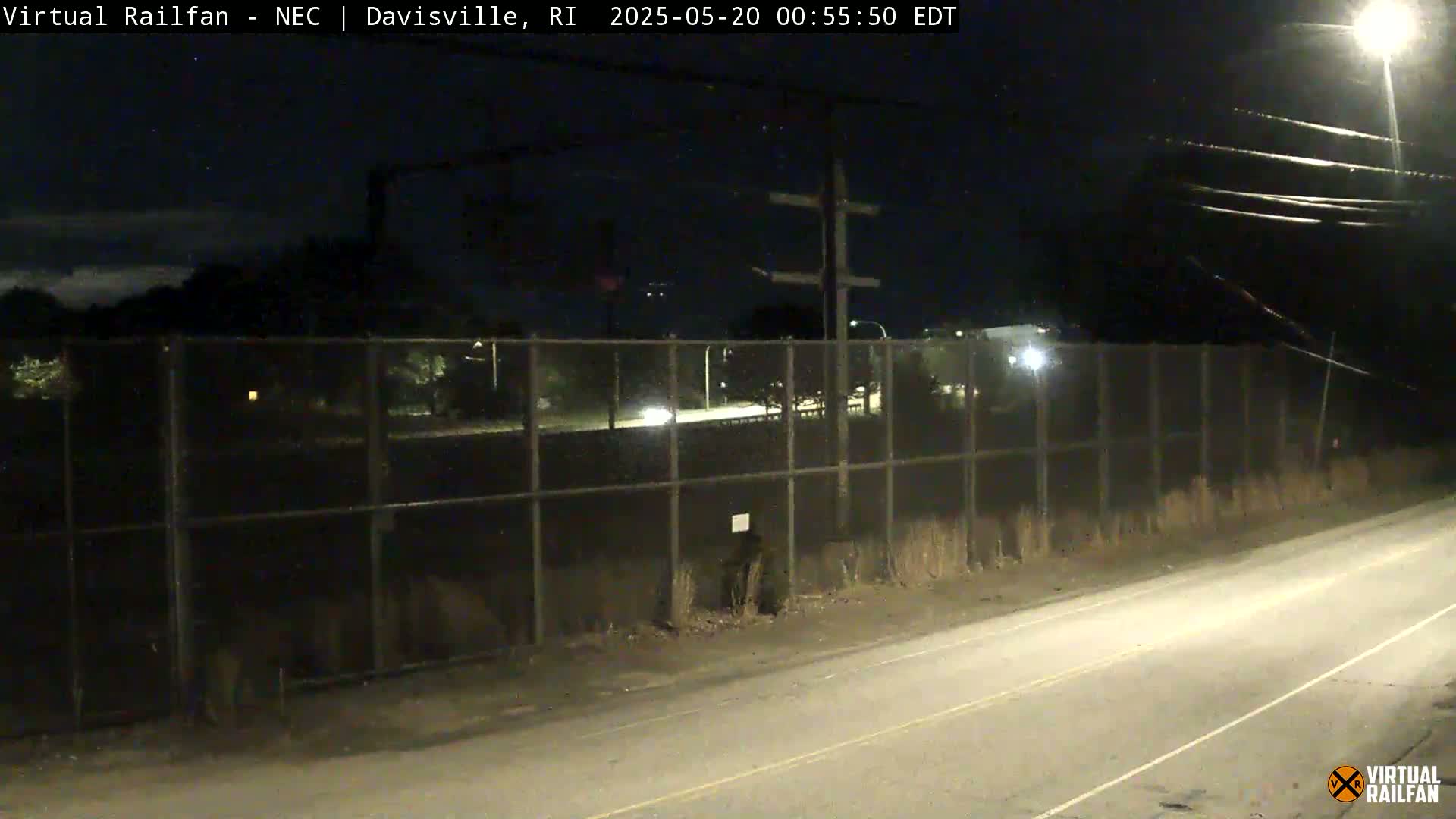 A nighttime view of a road next to a chain-link fence, beyond which are dimly lit trees and what appears to be a roadway with vehicles.