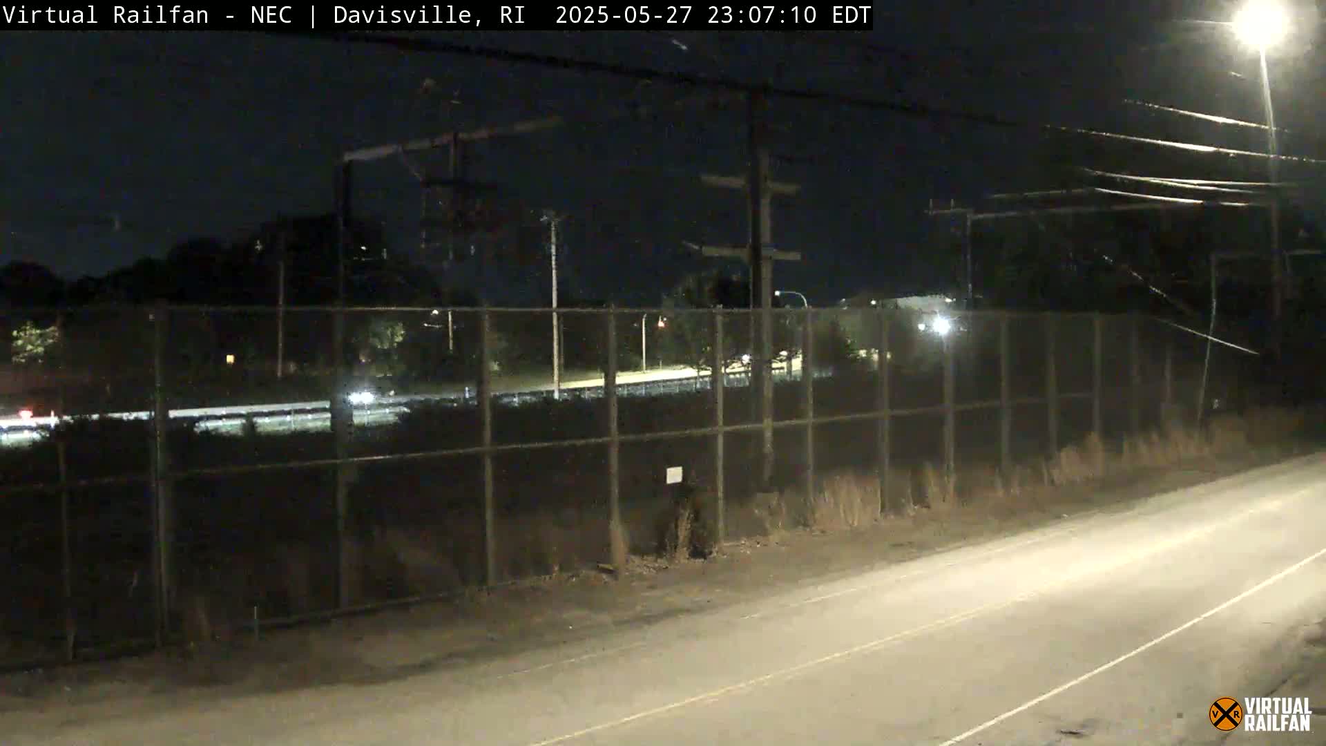 A nighttime view of a roadway next to a chain-link fence separating it from a railway line and some vegetation, under a dark sky.