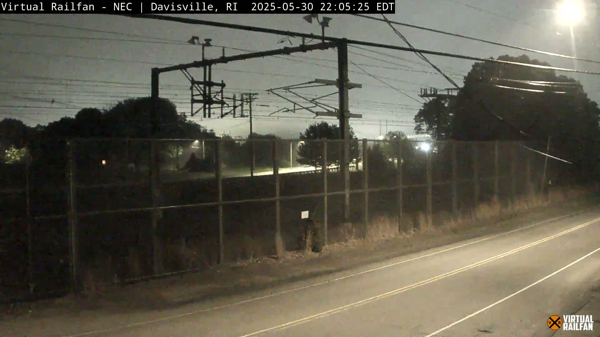 A nighttime view of a road beside a chain-link fence enclosing a rail yard under a dark, overcast sky.
