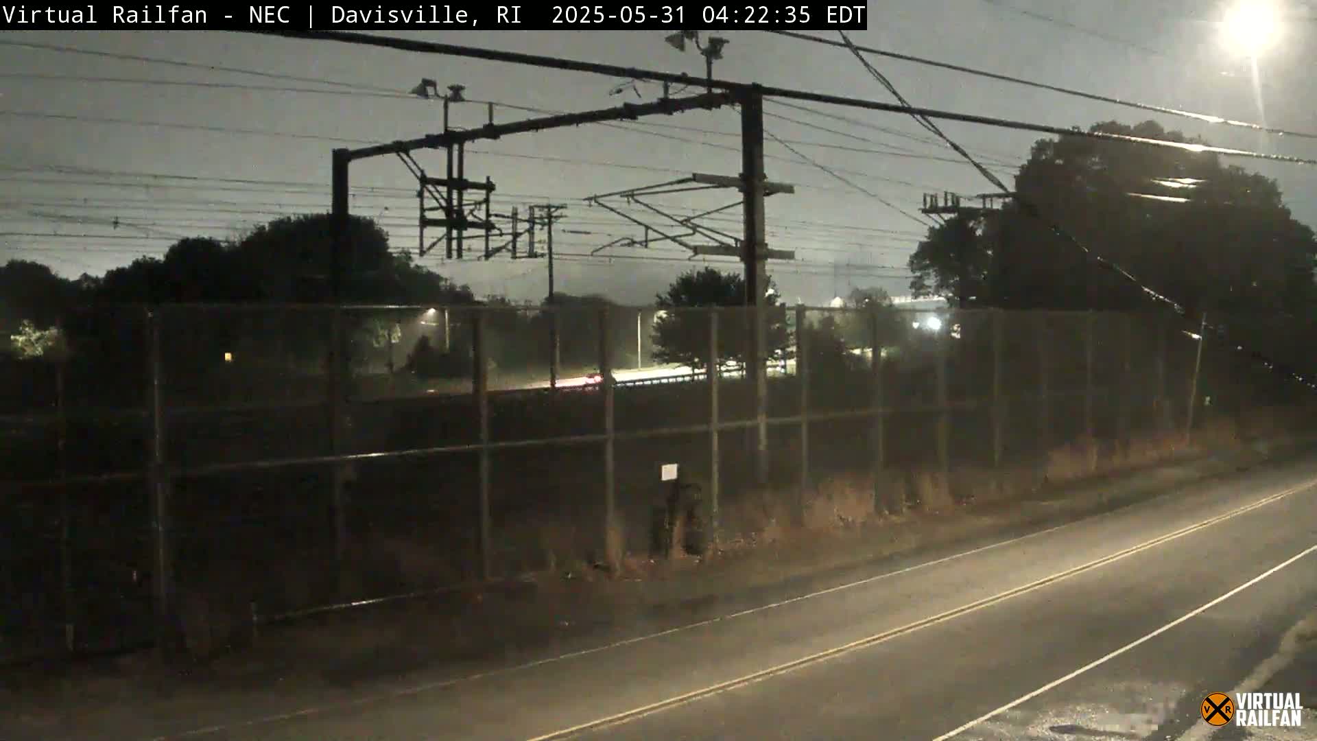 A nighttime view of a highway and railroad tracks behind a chain-link fence under a dark, possibly rainy sky.