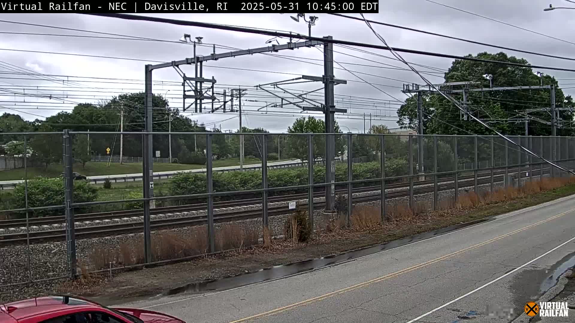 A view of railroad tracks behind a chain link fence on an overcast day, with a road and some vegetation in the foreground.