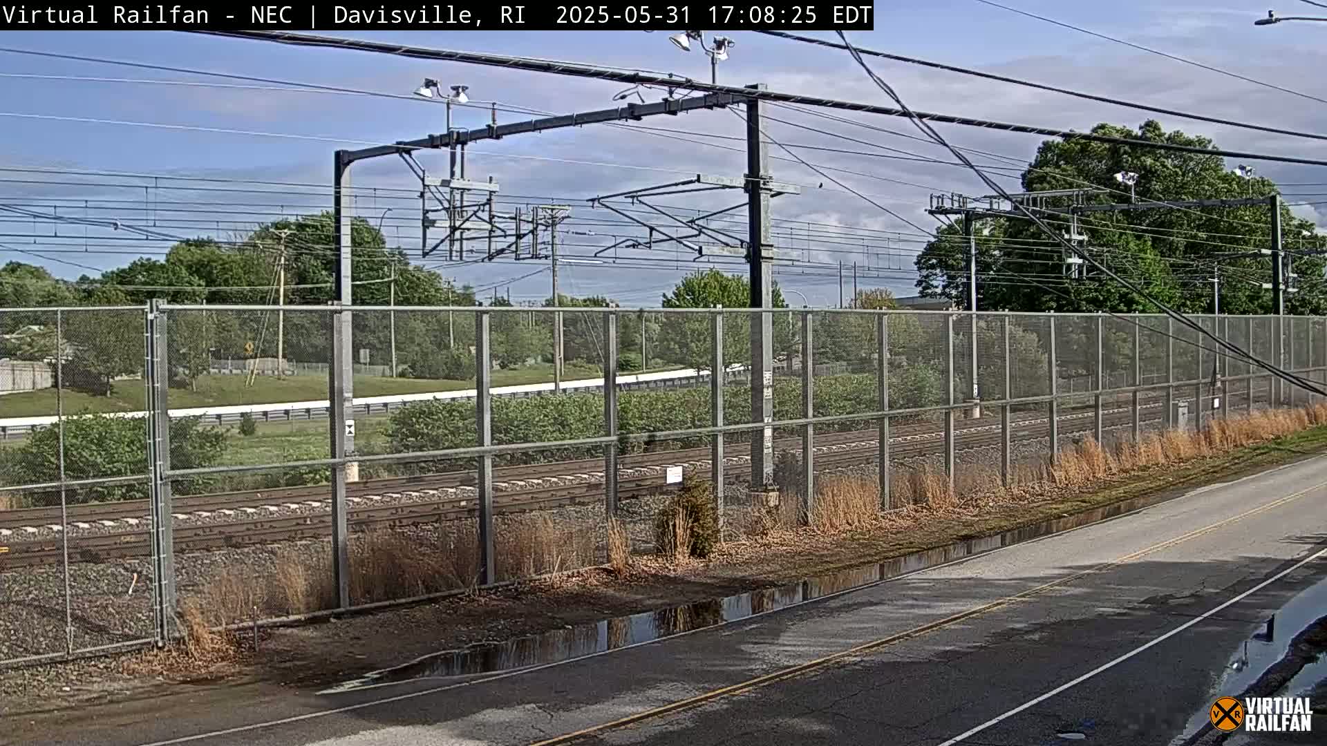 A mostly sunny outdoor scene shows a chain-link fence alongside railroad tracks, with some puddles on a nearby road.