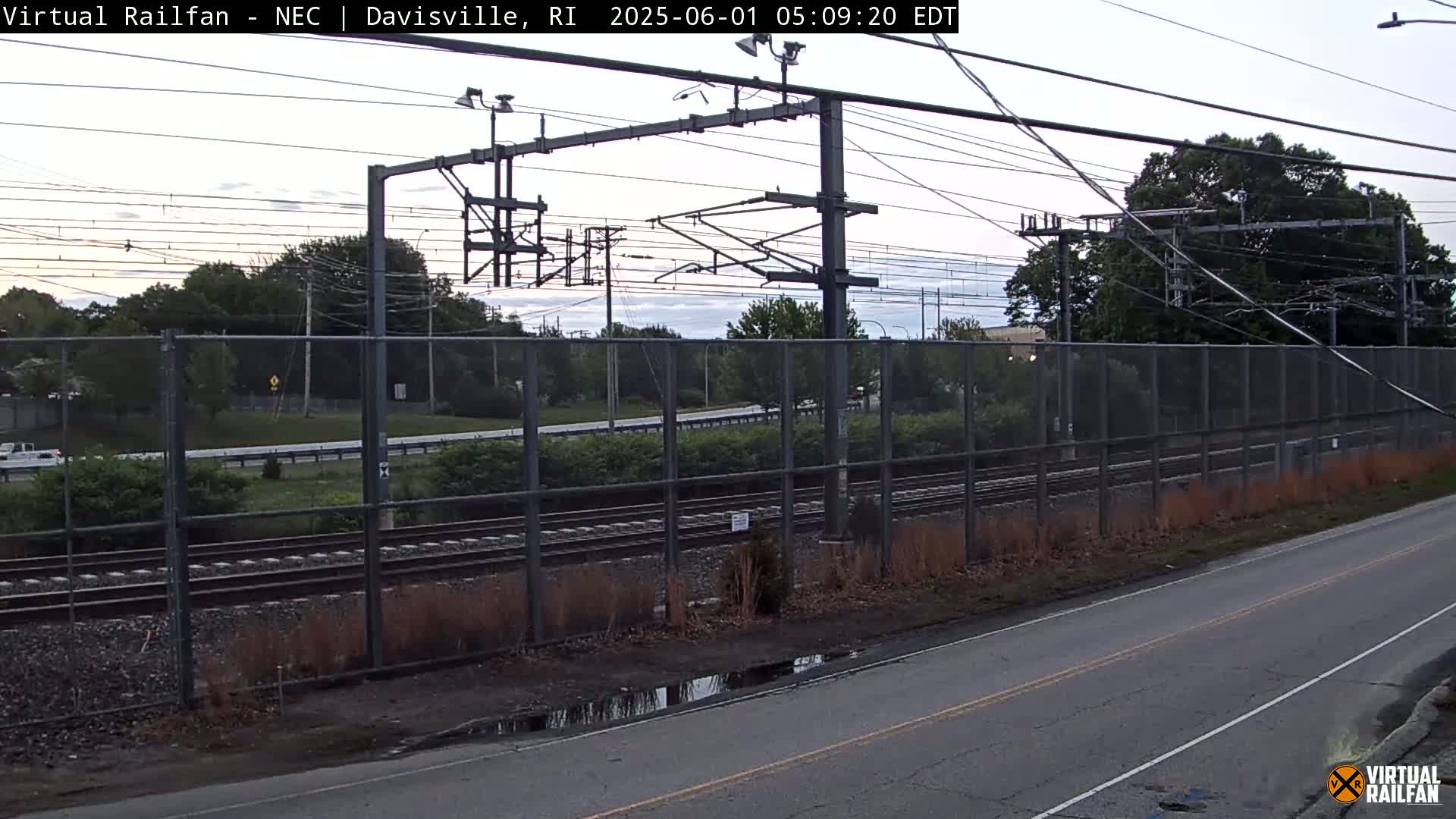 A roadway runs alongside a chain-link fence separating it from railroad tracks under an overcast sky.