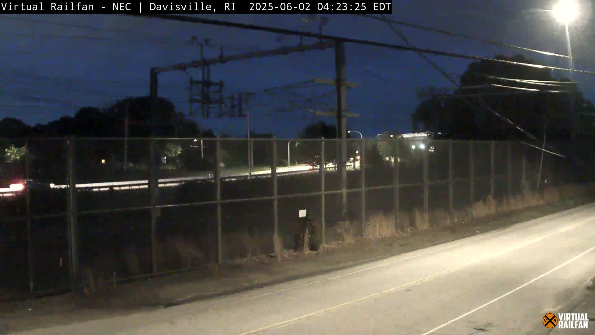 A nighttime view of a roadway beside a chain-link fence separating it from a railway, under a dark blue, clear sky.