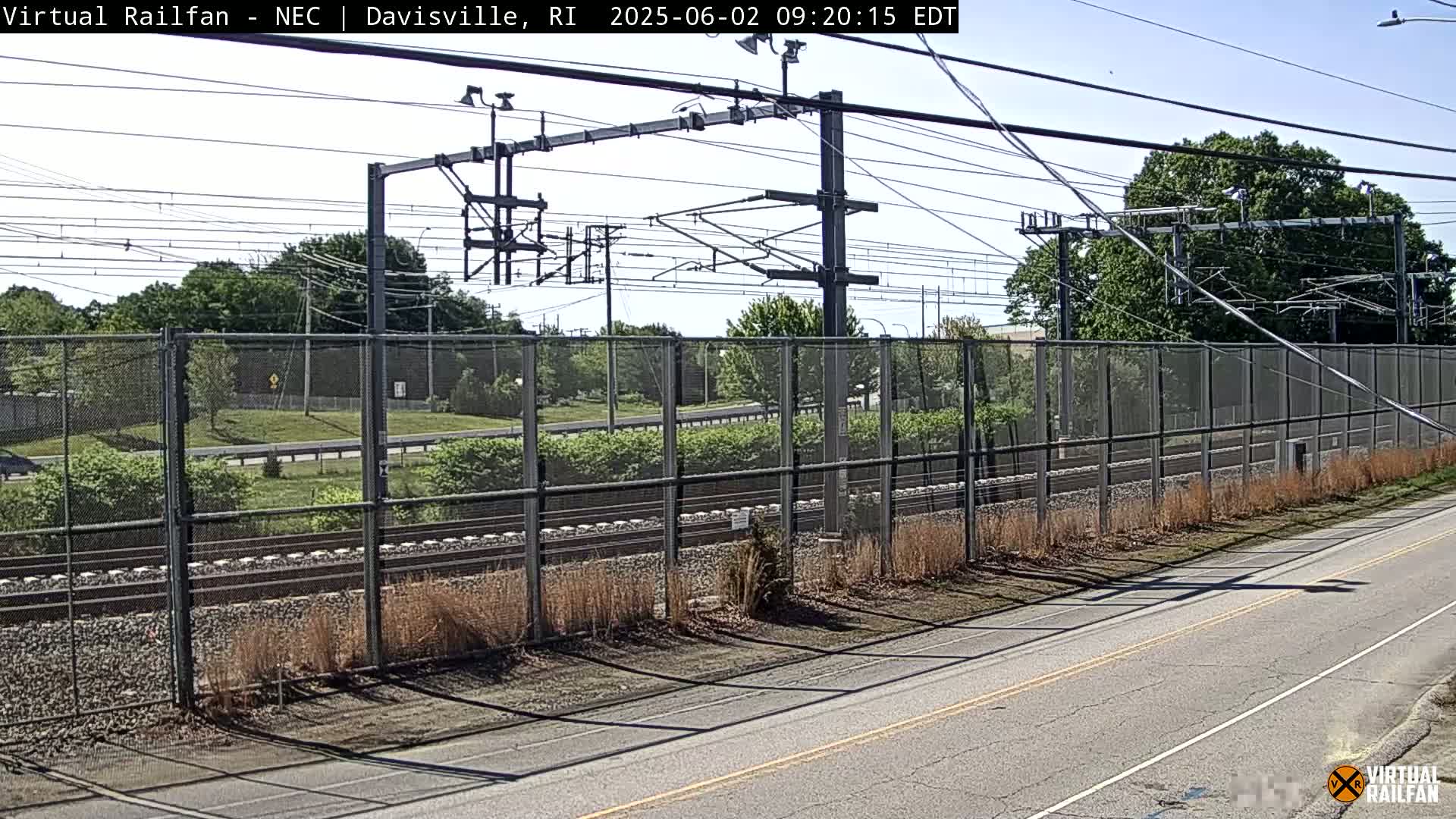 A sunny view of railroad tracks behind a chain-link fence beside a roadway, with overhead electrical lines and trees in the background.