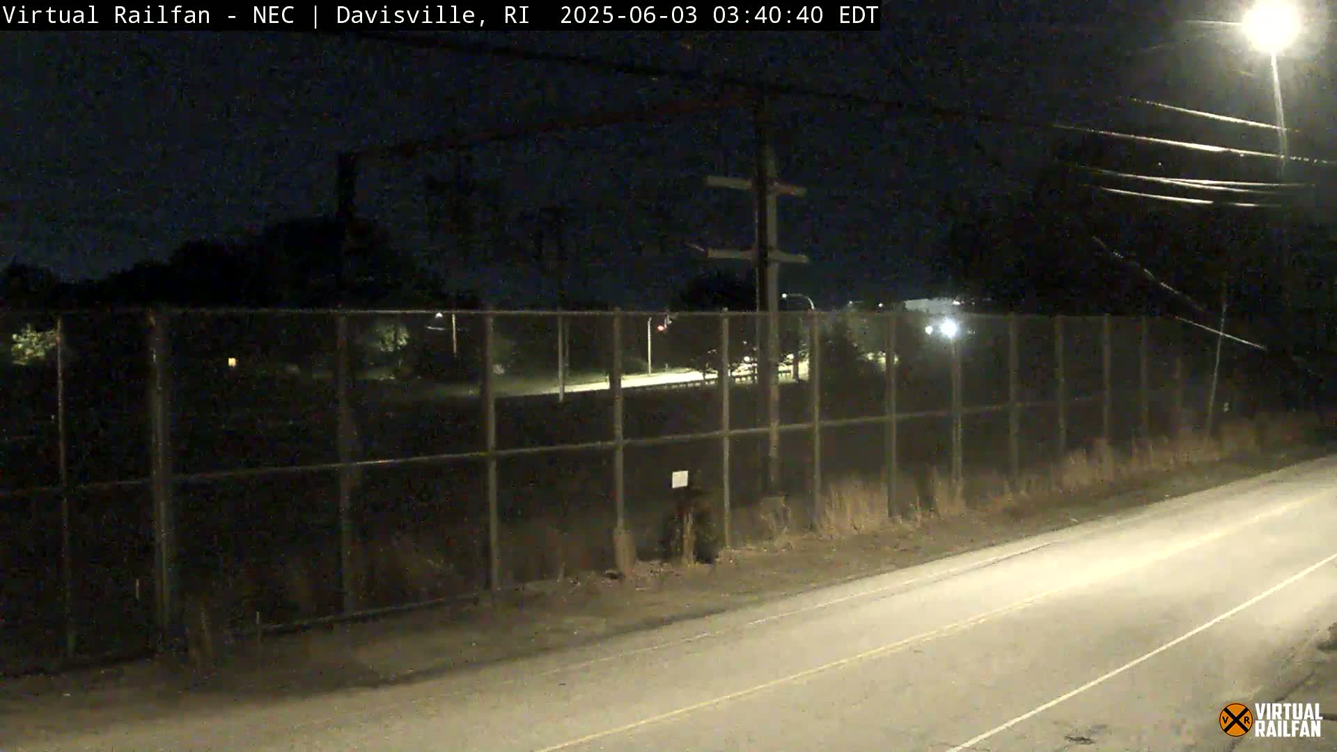 A nighttime view of a road next to a chain-link fence, beyond which are dimly lit trees and a roadway.