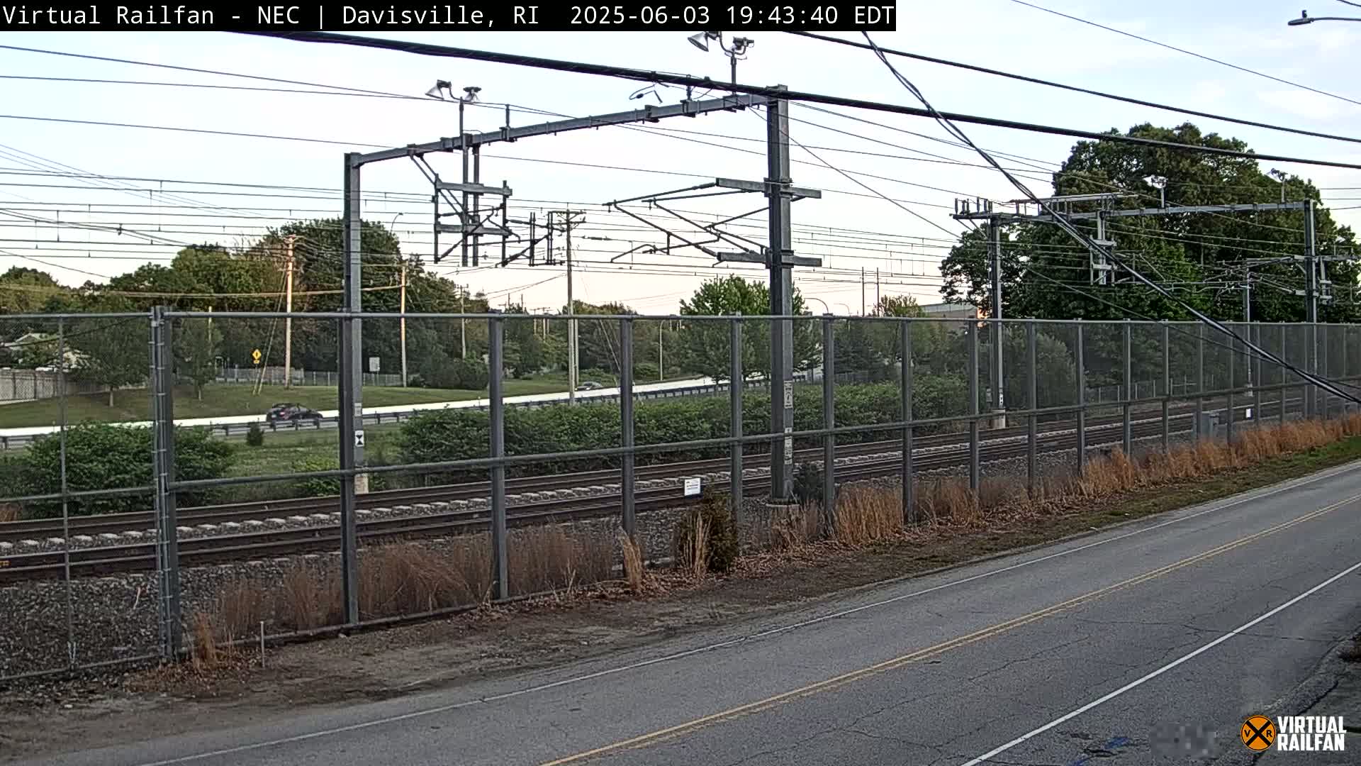 A view of railroad tracks behind a chain link fence, with a road and some cars visible beyond the fence under a partly cloudy sky.
