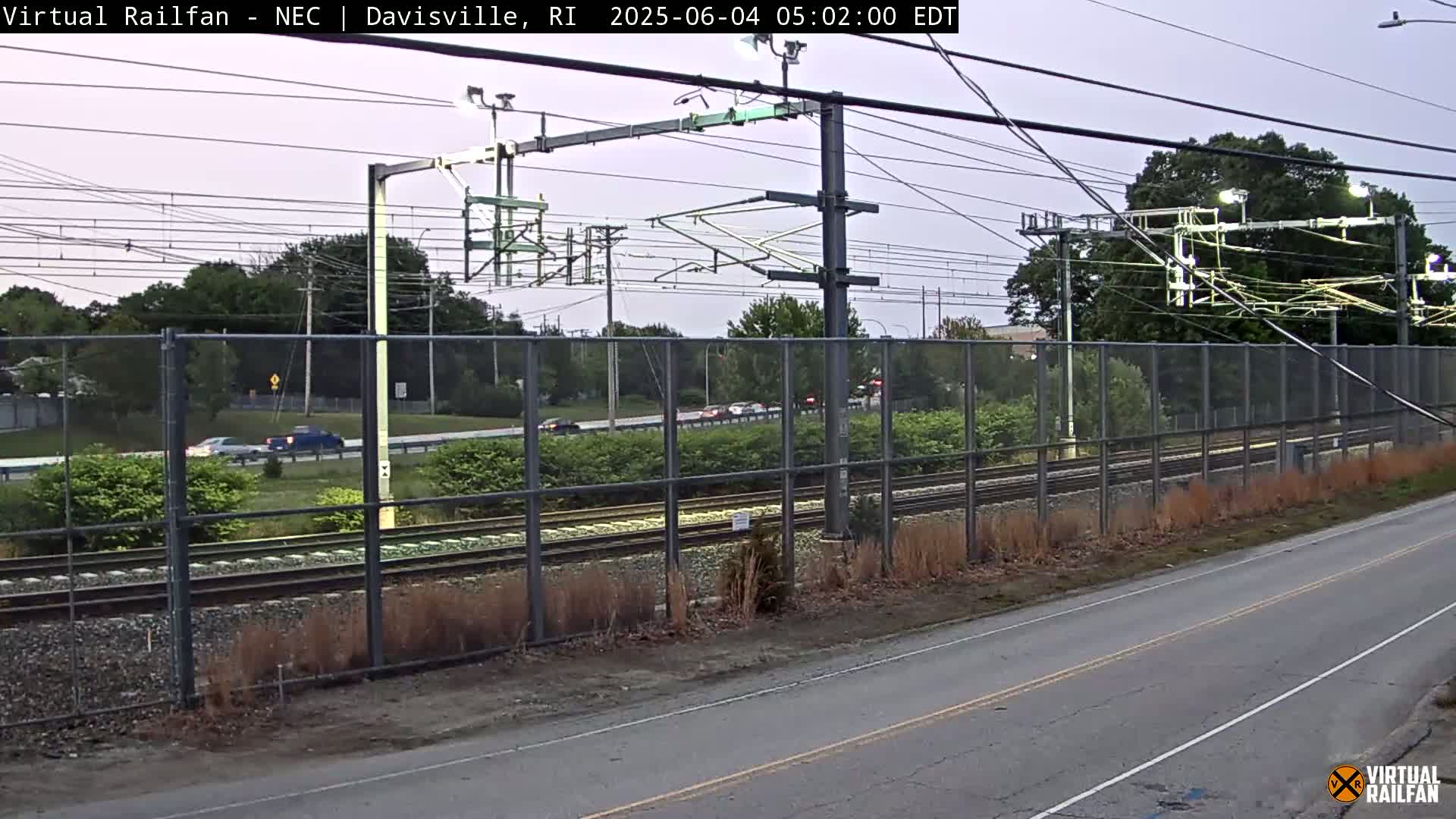 A view of a train track beside a road, separated by a chain-link fence, under an overcast sky.