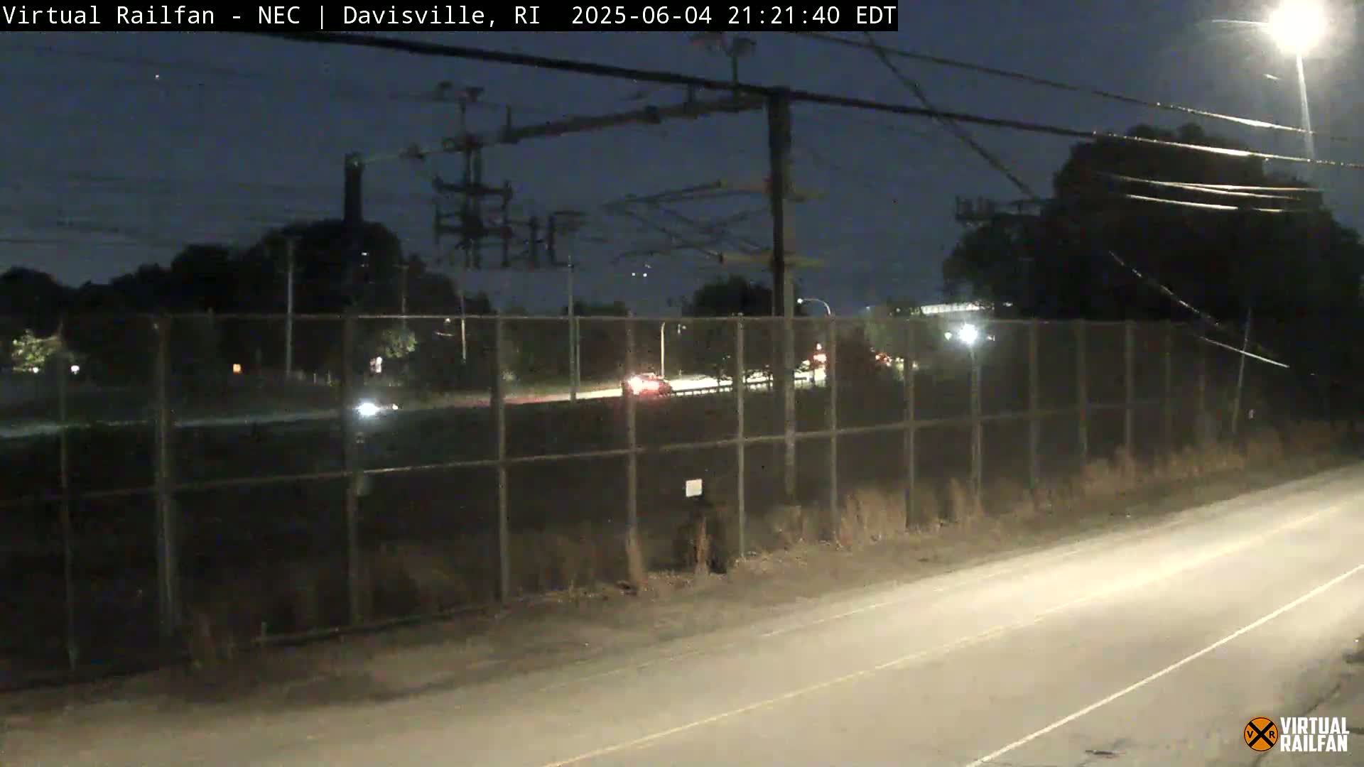 A nighttime view of a road beside a chain-link fence, beyond which are some trees and overhead power lines, with a few vehicles visible on the road.
