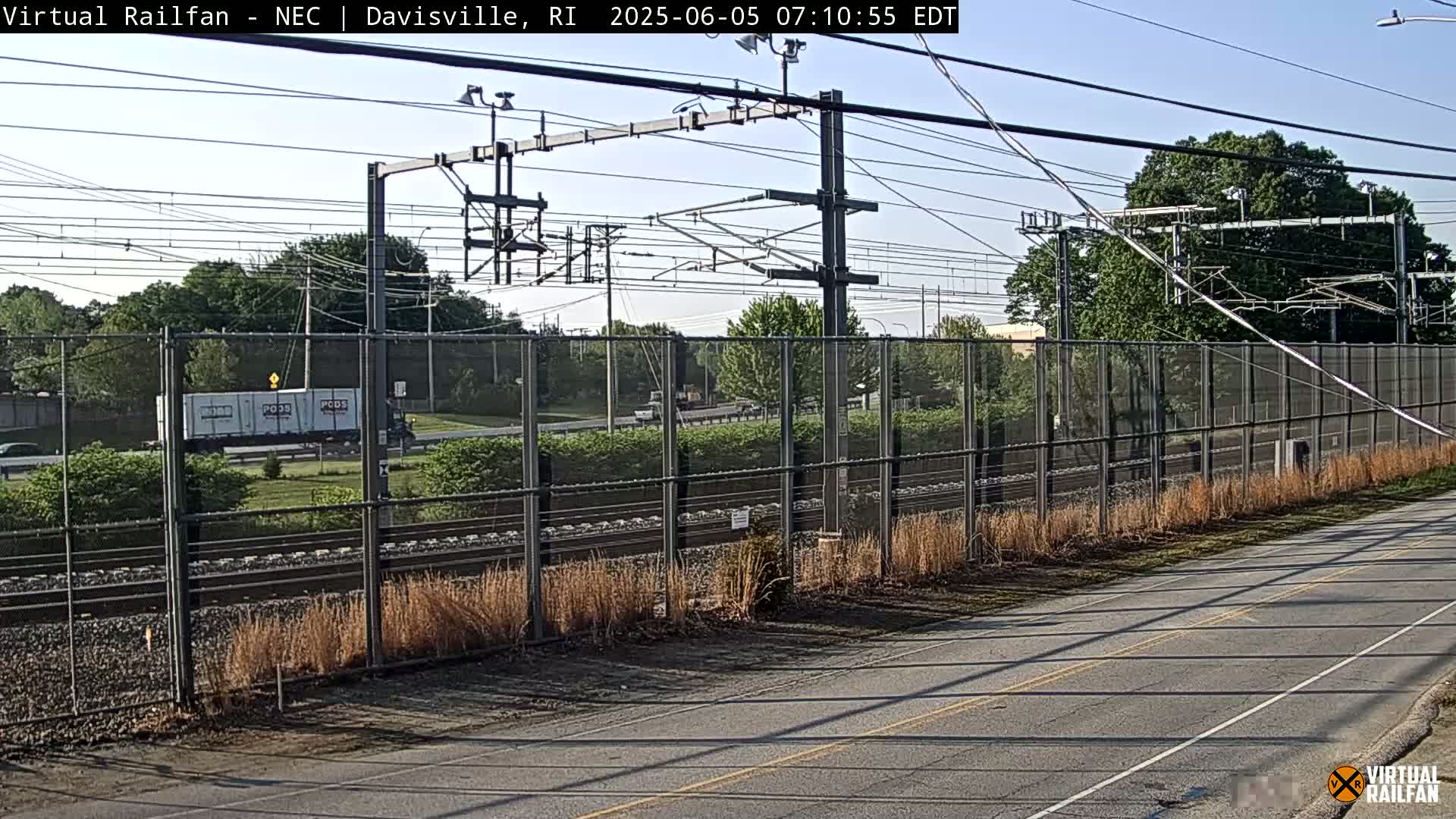 A road runs parallel to a railway track bordered by a chain-link fence and overgrown grass under a clear sky.