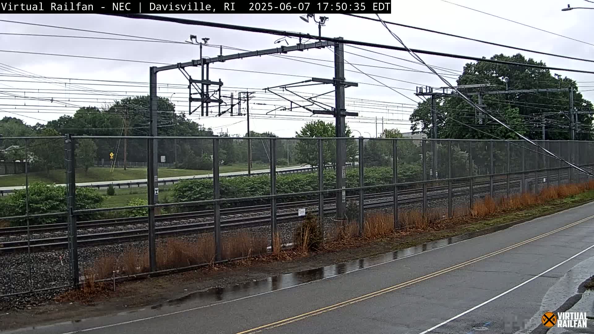 A view of railroad tracks behind a chain-link fence on an overcast day, next to a wet road.