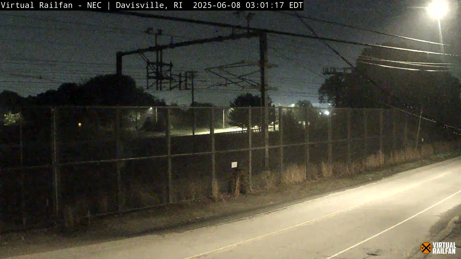 A nighttime view of a chain-link fence separating a road from an area with overhead electrical lines and trees under a dark sky.