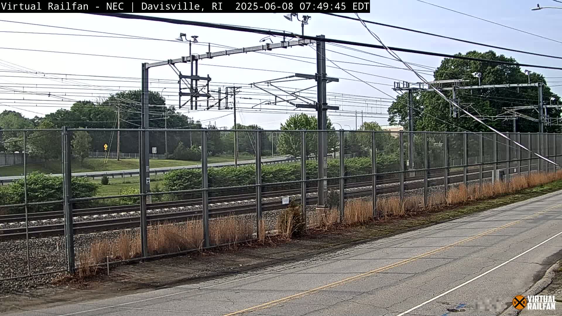 A view of train tracks beyond a chain-link fence on a partly sunny day, with a road in the foreground.
