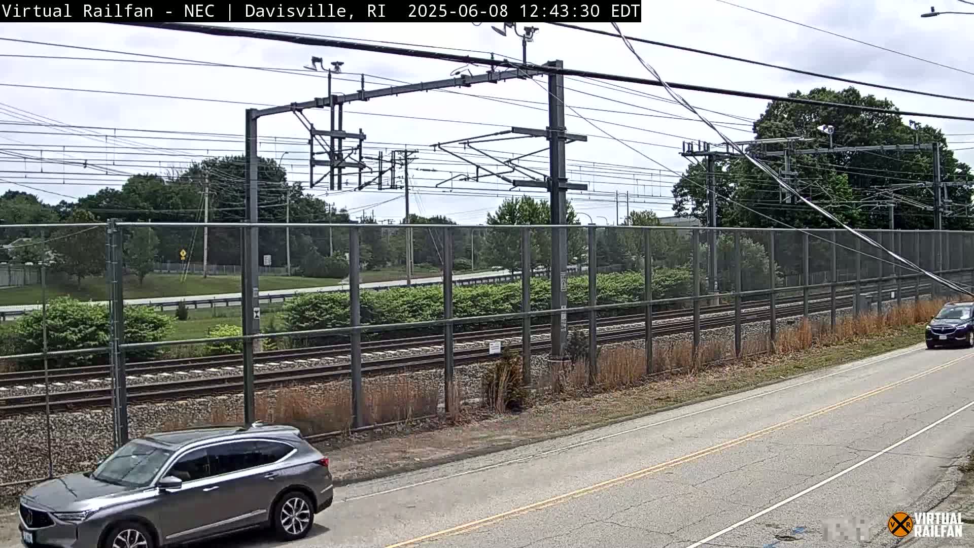 A partly cloudy day shows a view of train tracks beside a road, with a gray SUV parked near the road and a dark-colored SUV driving away.